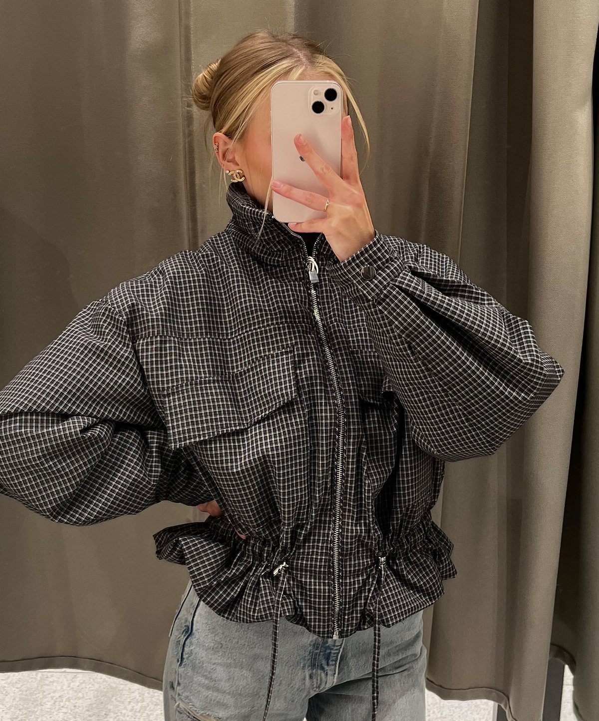 Woman in a checkered puff-sleeve jacket and light blue jeans taking a mirror selfie.