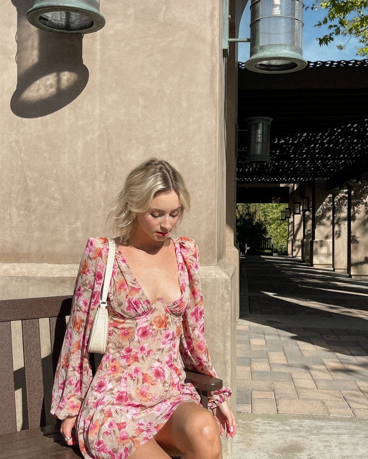 A blonde woman wearing a pink floral long-sleeve mini dress sitting on a wooden bench.
