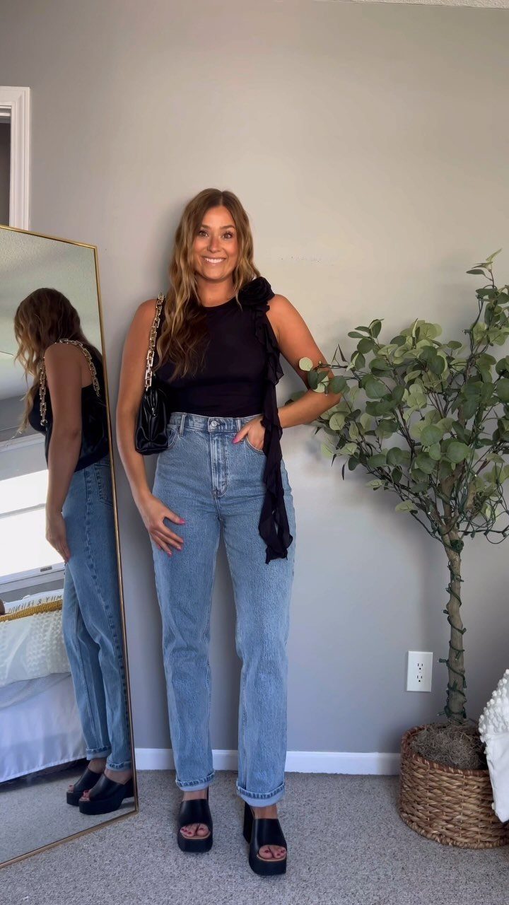 A smiling woman posing in a black top, light blue jeans, and black platform sandals.