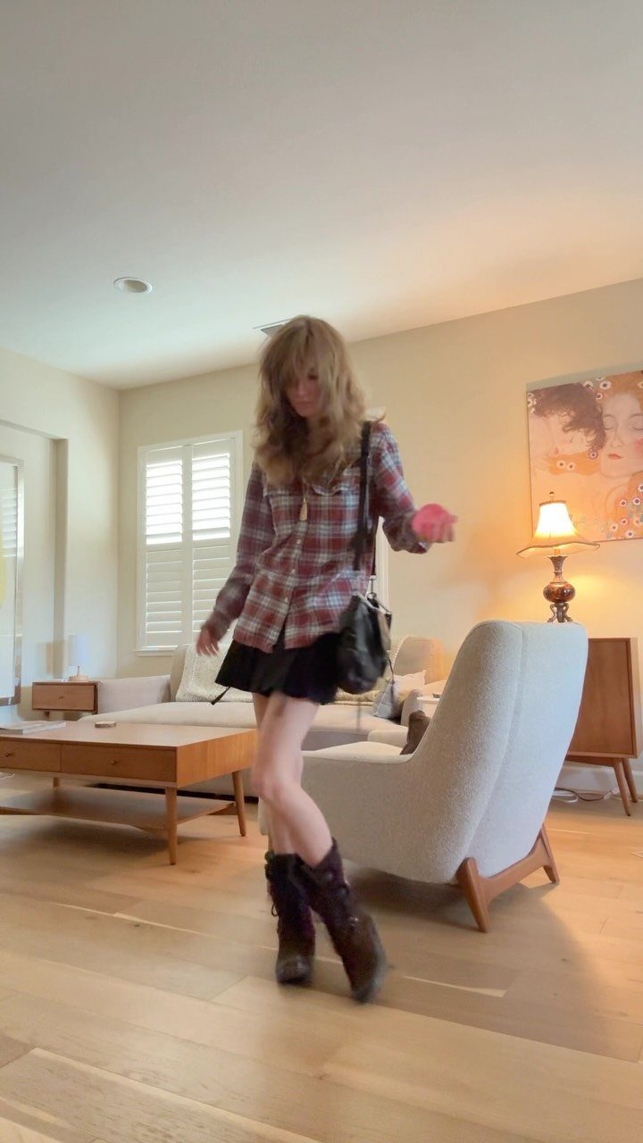 A person wearing a plaid flannel shirt, black skirt, and boots in a living room.
