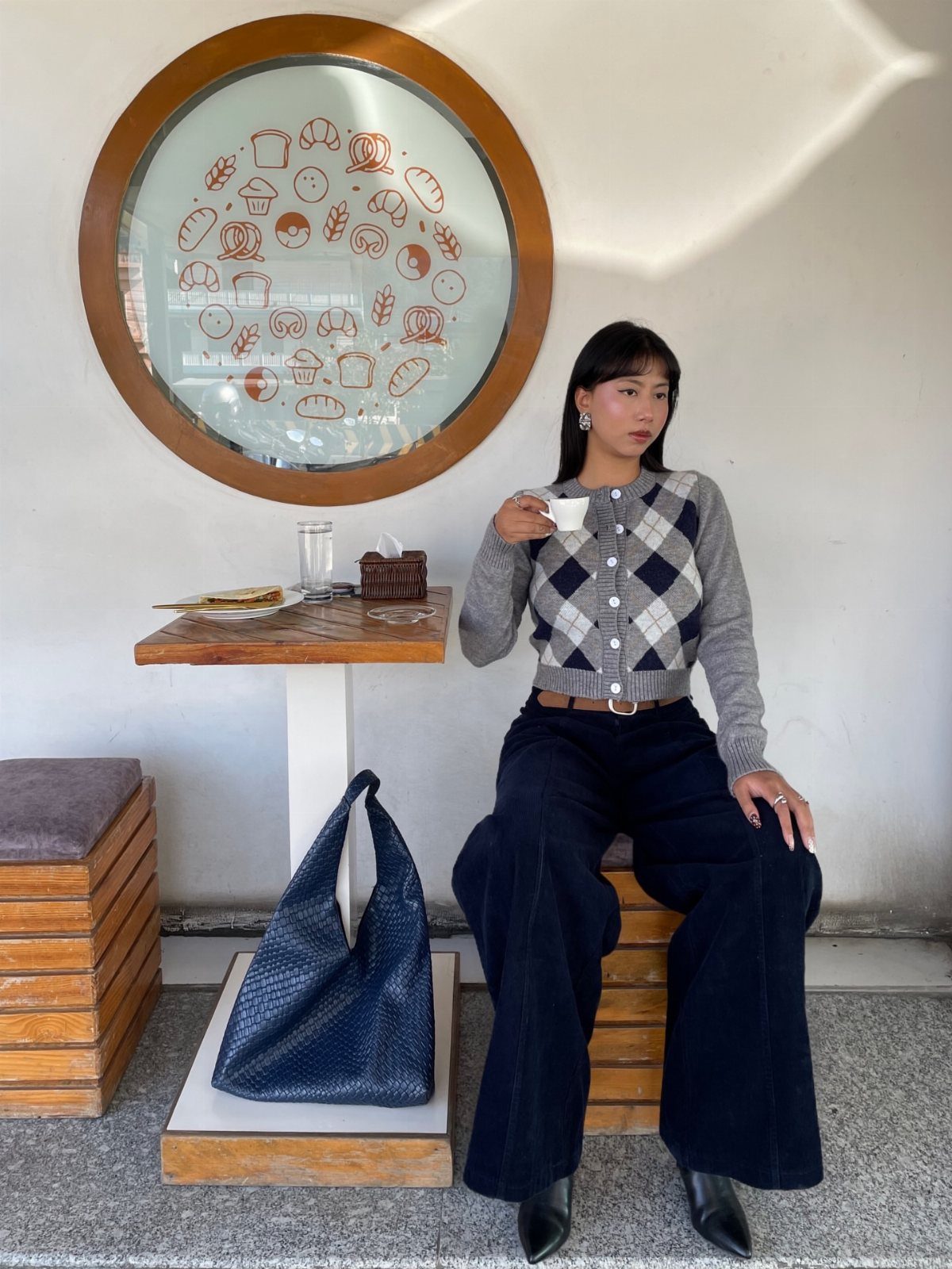 A woman wearing dark high-waisted wide-leg trousers and a patterned cardigan sitting by a cafe table.