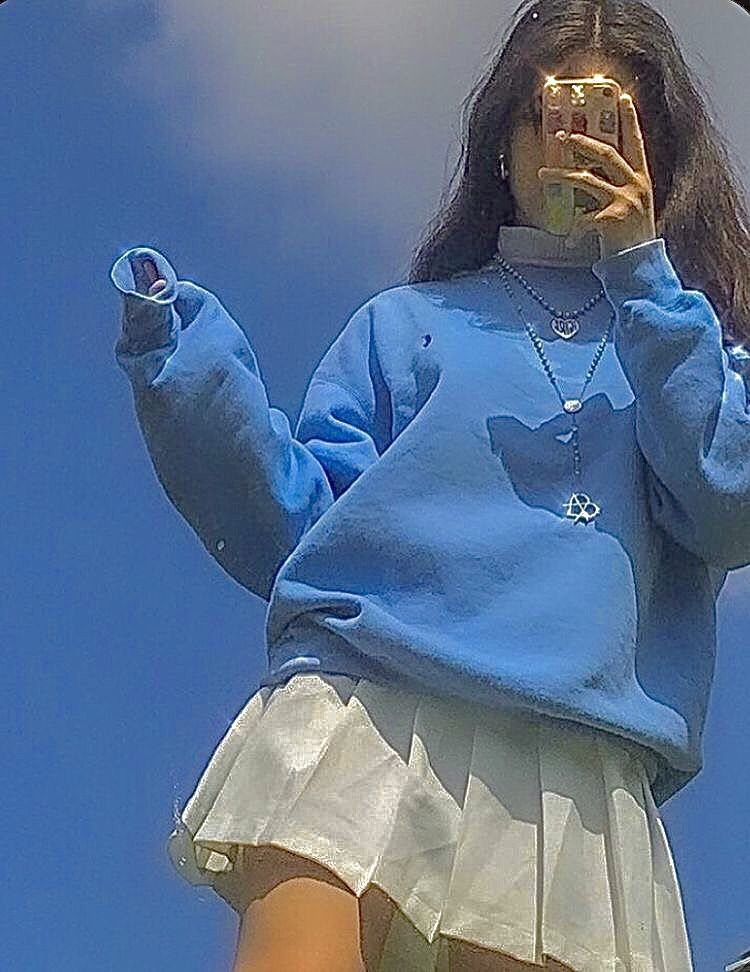 Person in a blue sweatshirt and white pleated skirt wearing layered silver chain necklaces.
