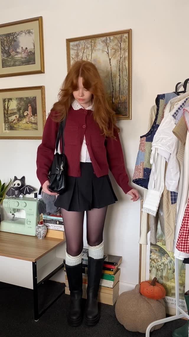 A young woman with red hair wearing a maroon cardigan, black pleated skirt, and black boots.