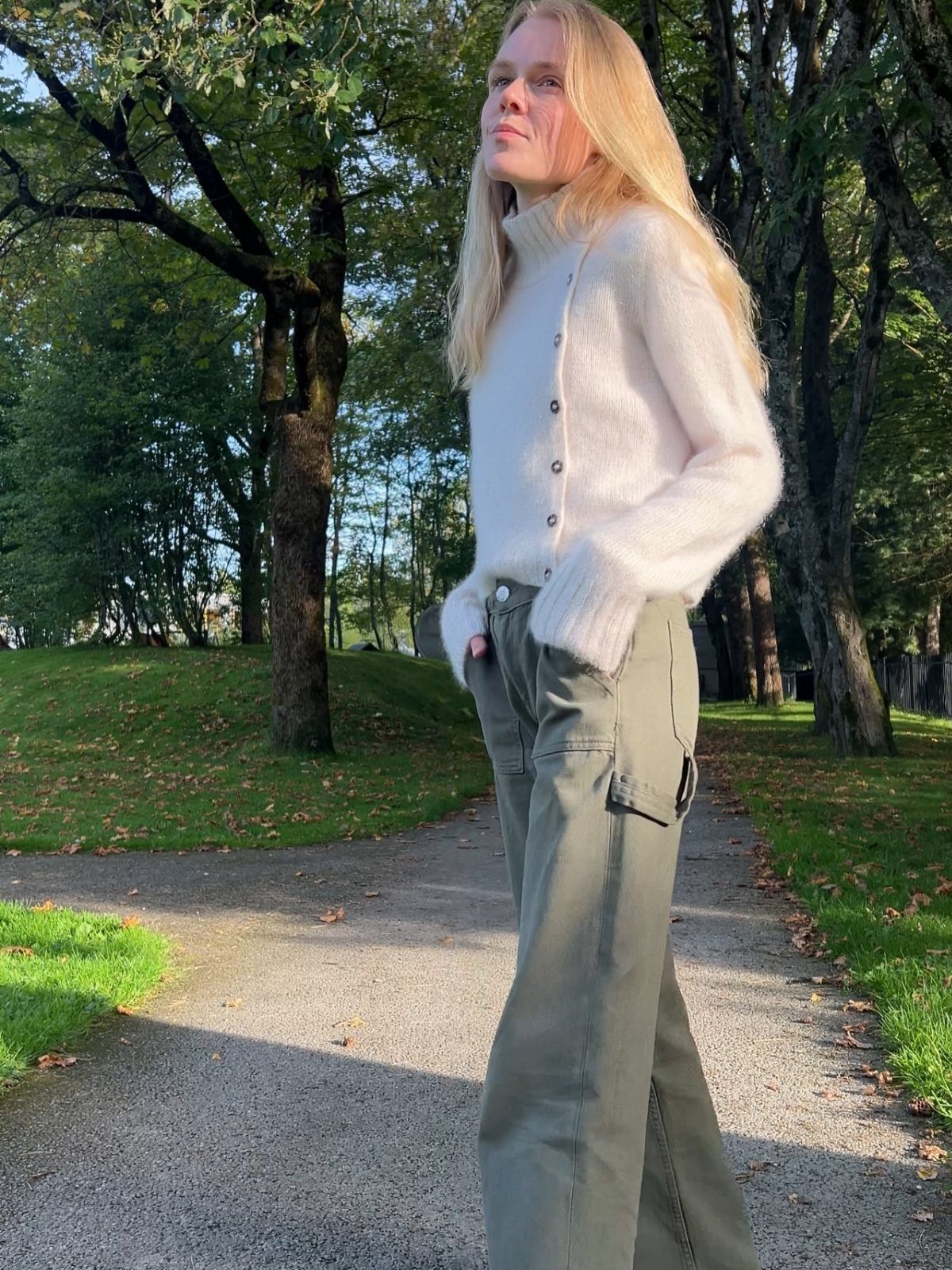 A blonde woman wearing a white fuzzy cardigan and olive green cargo pants standing outdoors.