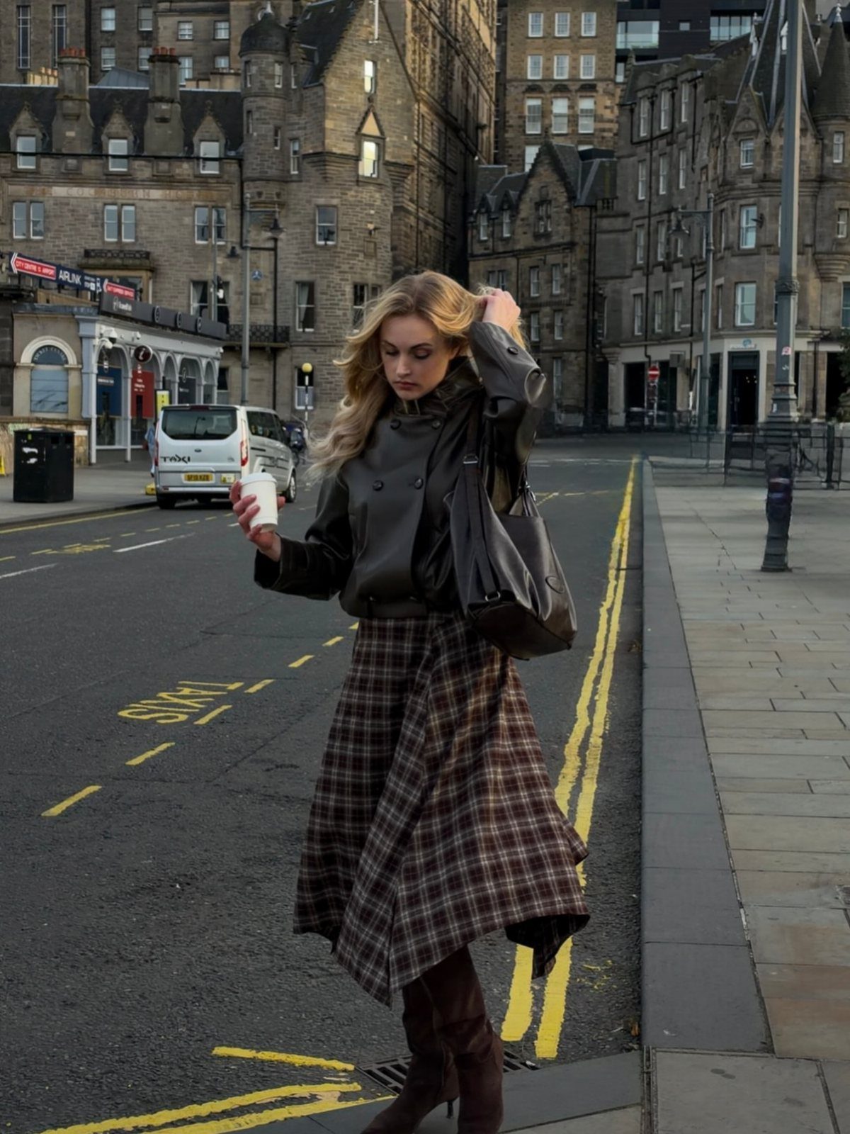 A woman in a leather jacket and long plaid skirt walking down a city street.