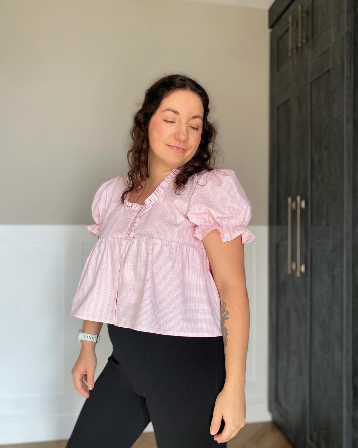 A woman wearing a pink gingham puff-sleeve blouse and black leggings standing indoors.