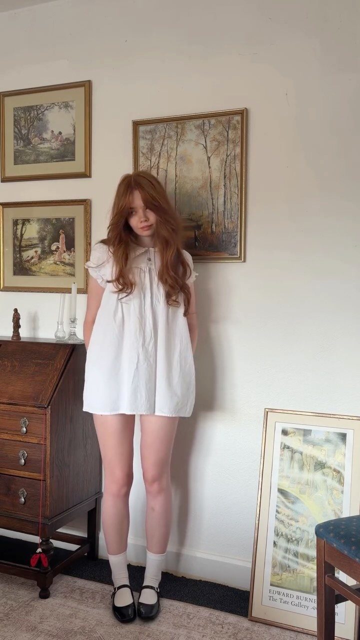 A young woman with red hair wearing a white smock dress and black Mary Jane shoes.