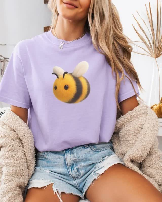 Woman wearing a lavender bumblebee graphic t-shirt and denim shorts sitting outdoors.