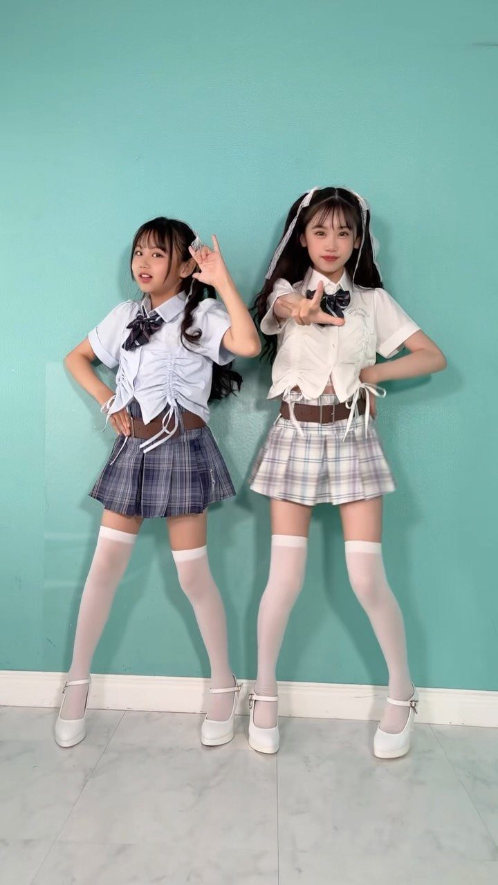 Two girls posing in plaid skirts, white shirts, and knee-high socks against a teal wall.