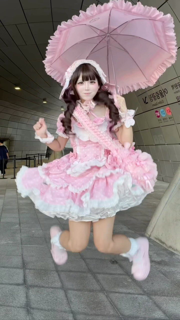 Woman in a pink ruffled dress jumping while holding a pink parasol and wearing sneakers.