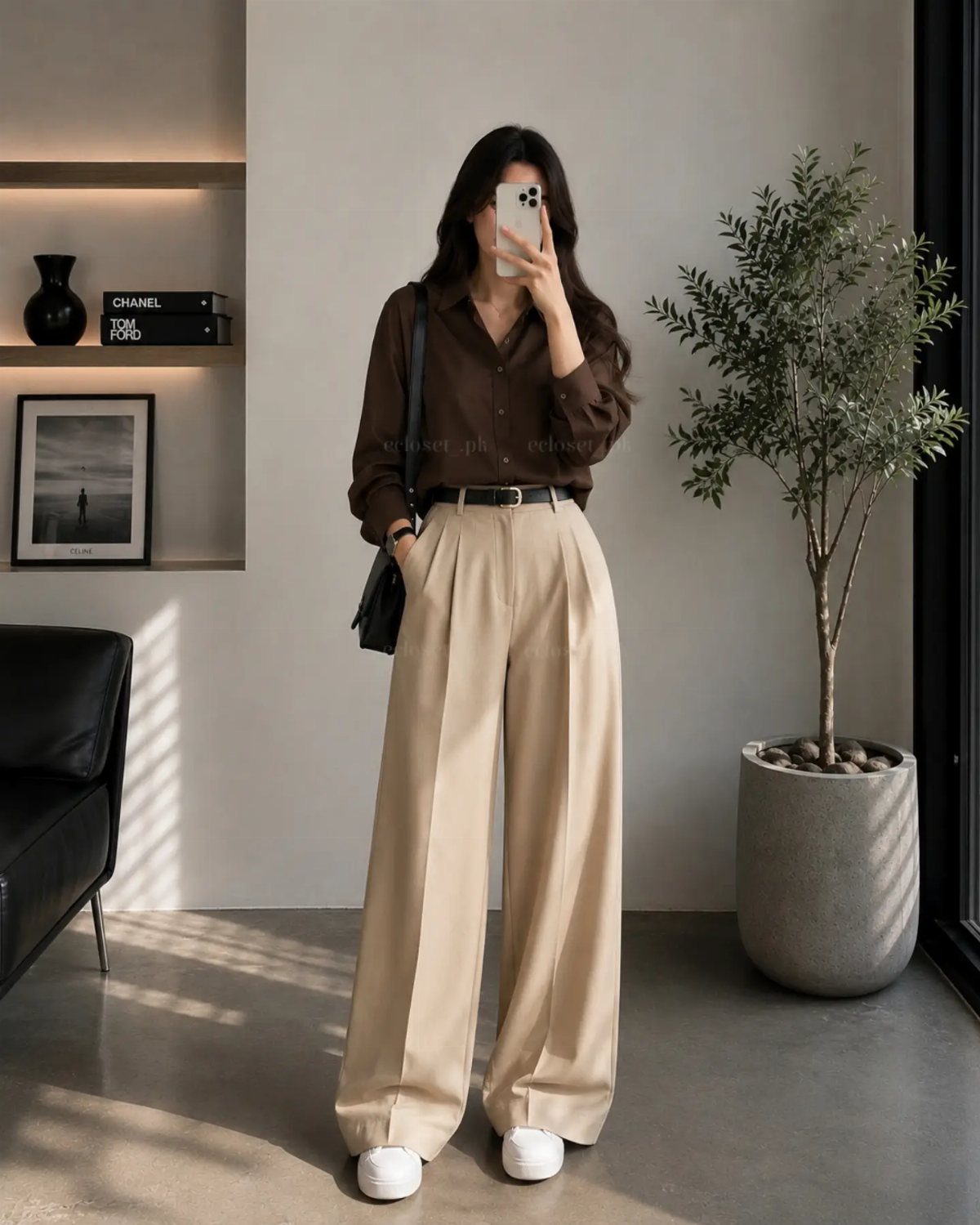 A woman posing in a brown shirt and wide-leg beige trousers for a mirror selfie.