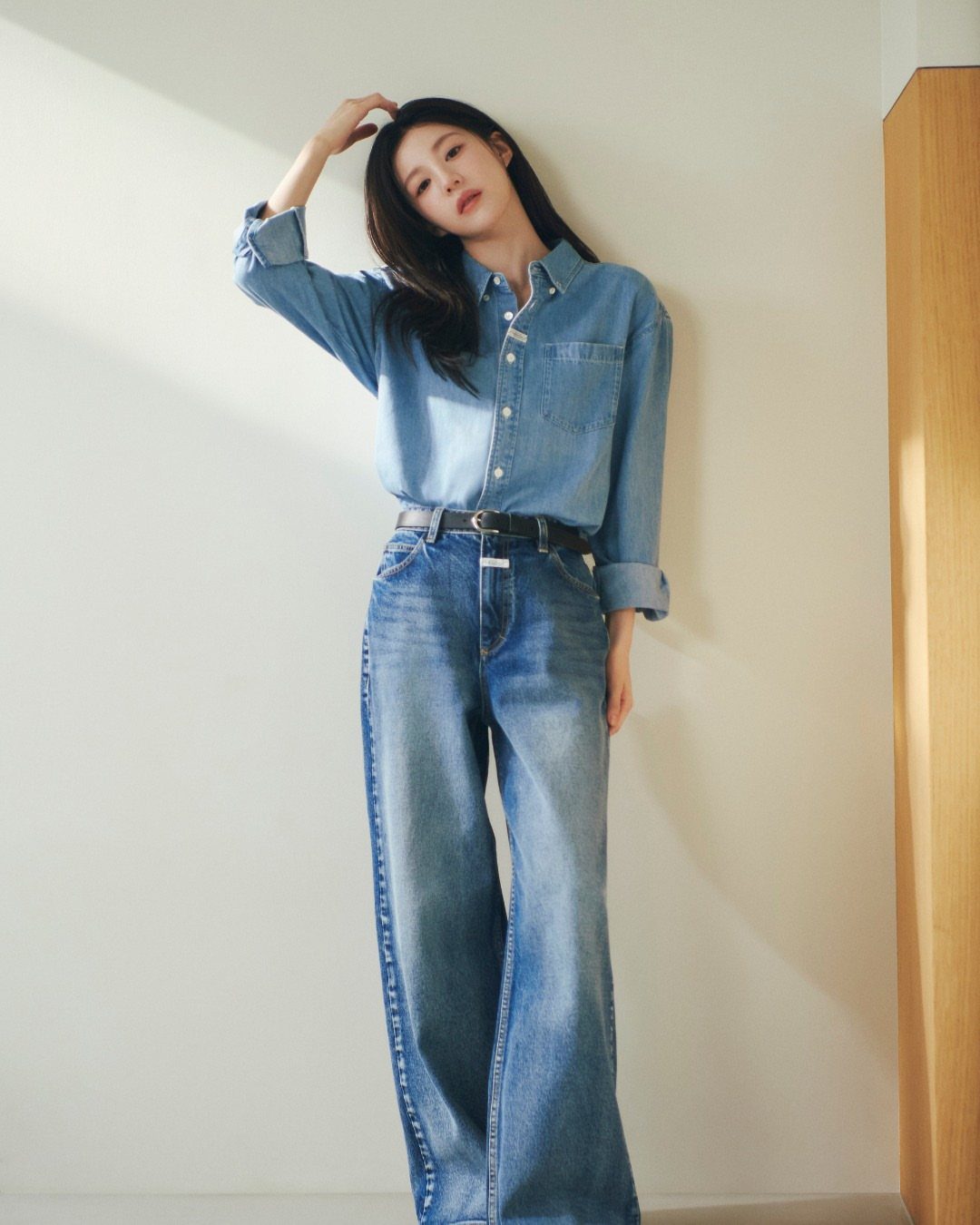 A woman wearing a denim shirt and high-waisted wide-leg jeans posing against a white wall.