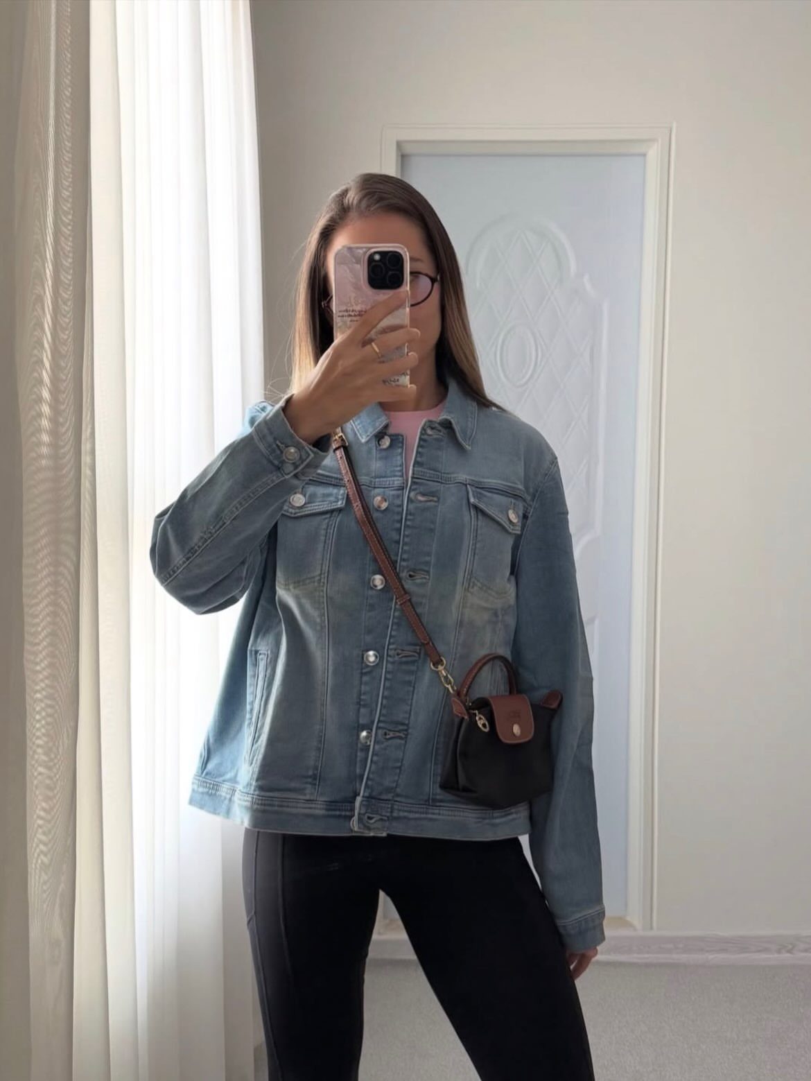 Woman in a denim jacket and black pants taking a mirror selfie indoors.