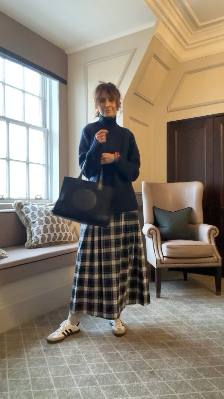 A woman posing in a navy turtleneck, plaid maxi skirt, and white sneakers while holding a black tote.