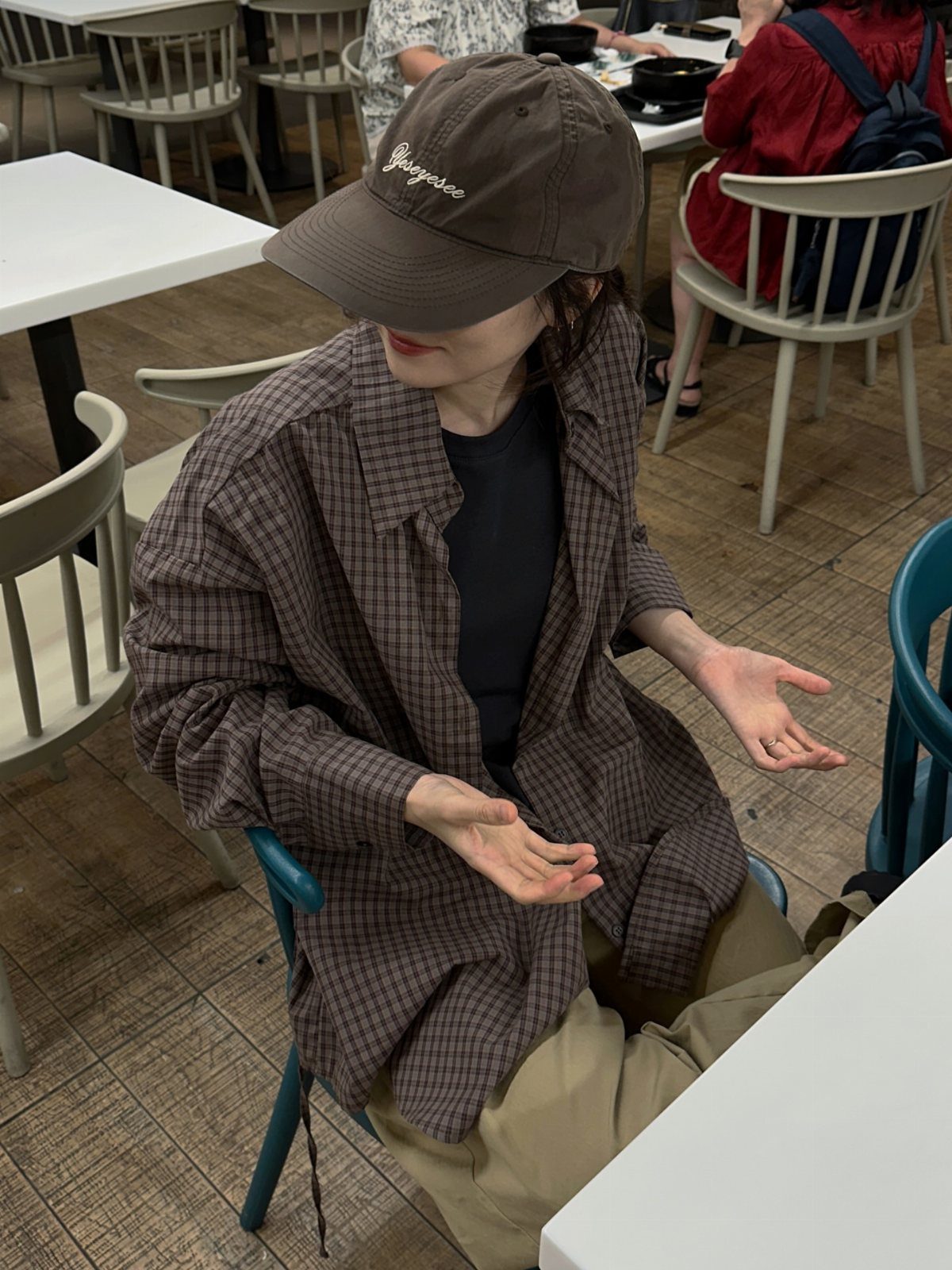A person wearing an oversized checkered shirt and a brown baseball cap sitting at a cafe table.