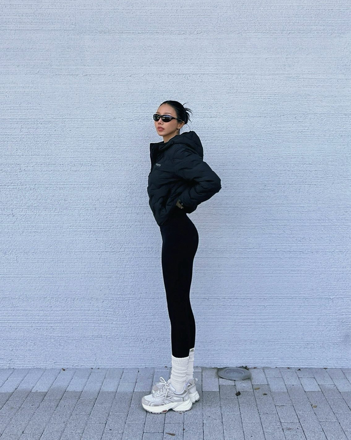 Woman in black puffer jacket, black leggings, and chunky sneakers posing against a grey wall.