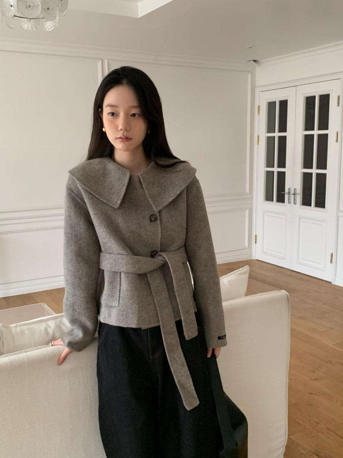 A young woman wearing a grey belted wool jacket and a black long skirt indoors.
