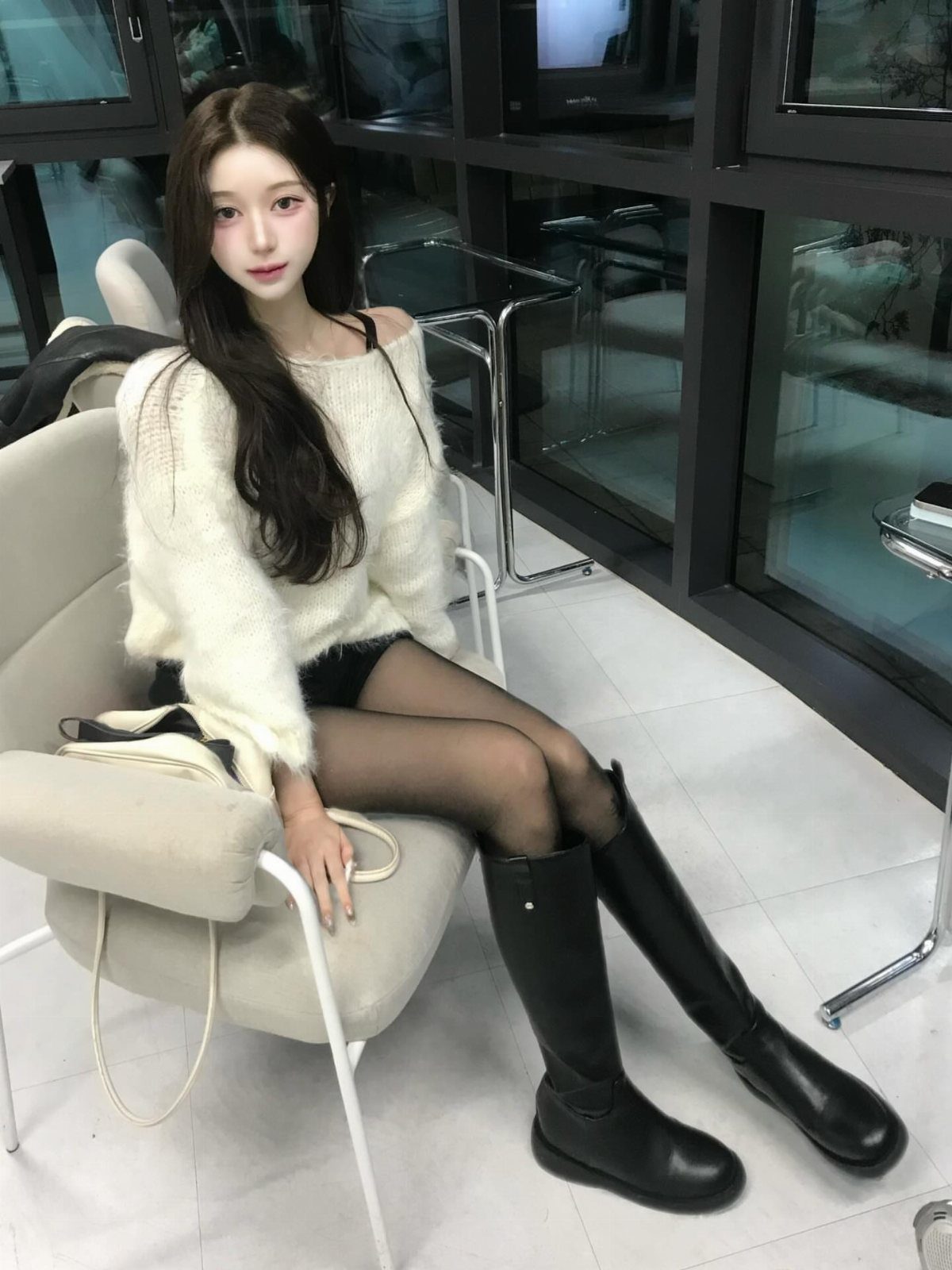 A woman wearing a white fuzzy sweater, black tights, and tall black leather boots sitting in a chair.