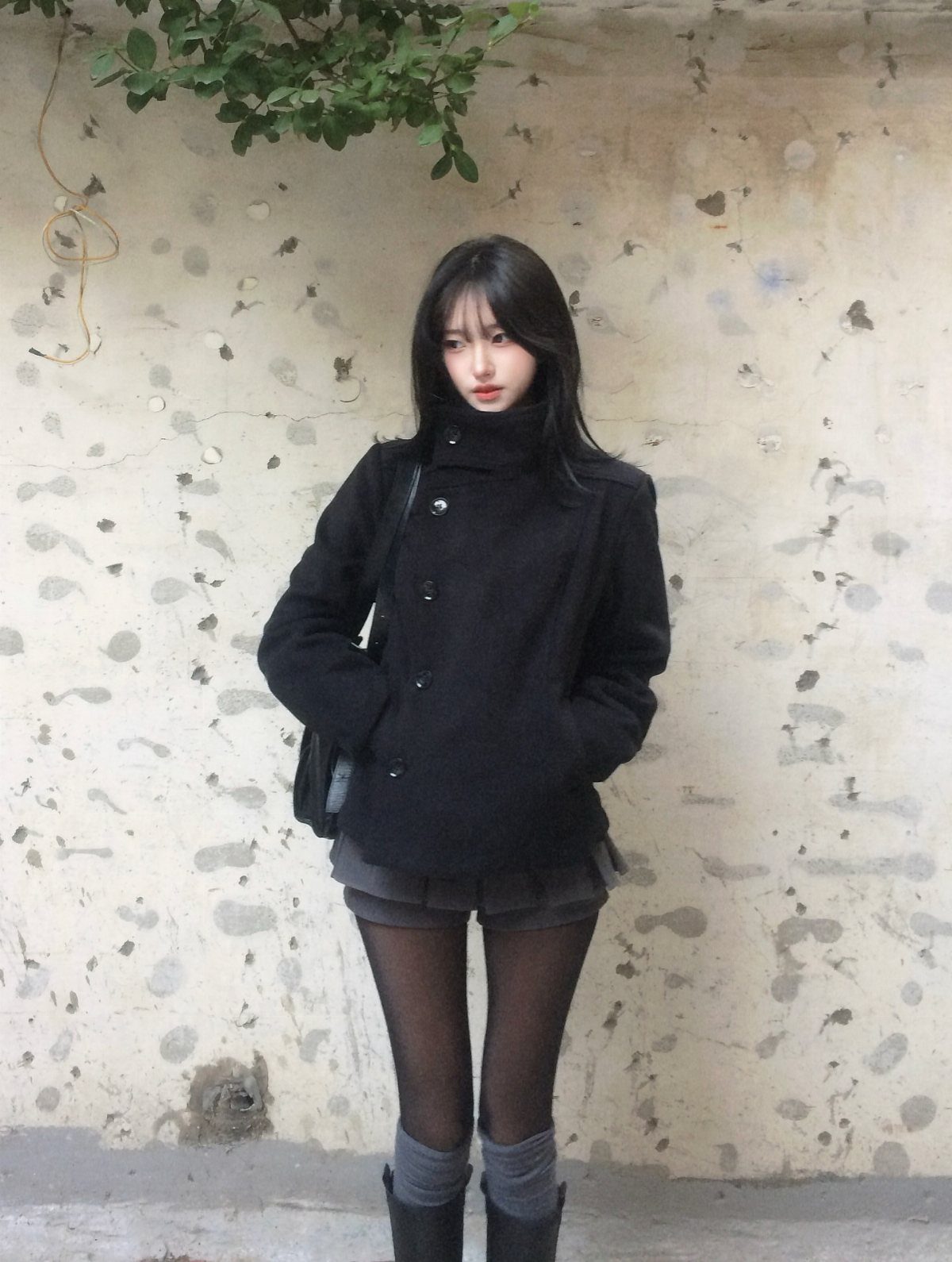 A young woman posing in a black coat, grey skirt, and black tights against a wall.