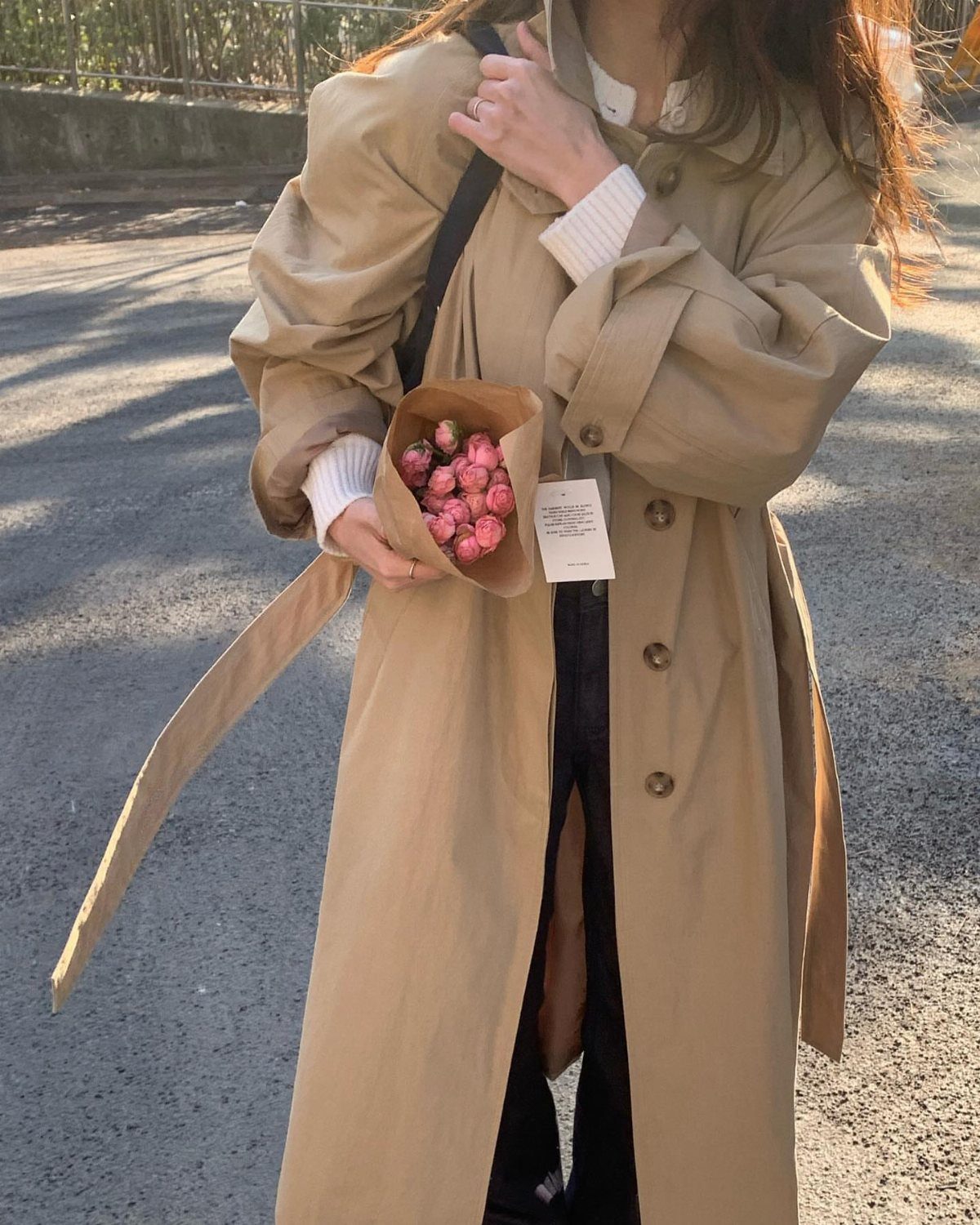A person wearing an oversized tan trench coat while holding a small bouquet of pink flowers.