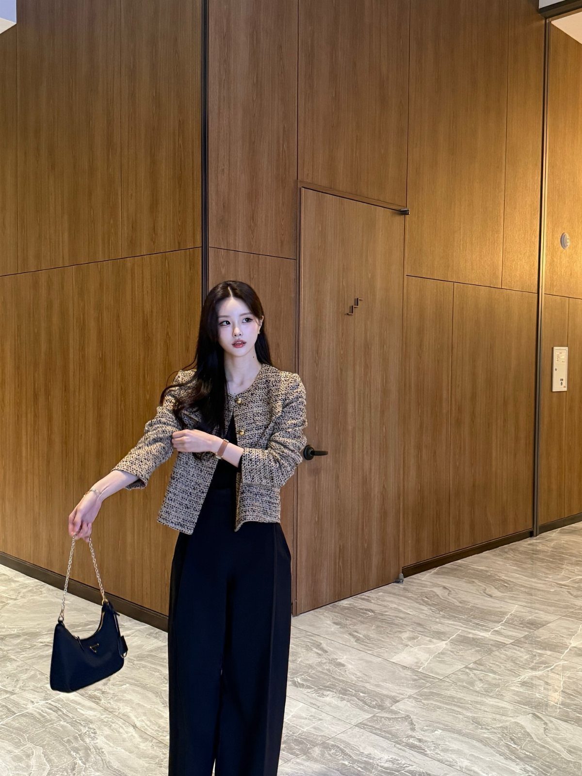 Woman wearing a textured tweed jacket and black wide-leg trousers standing against a wooden wall.