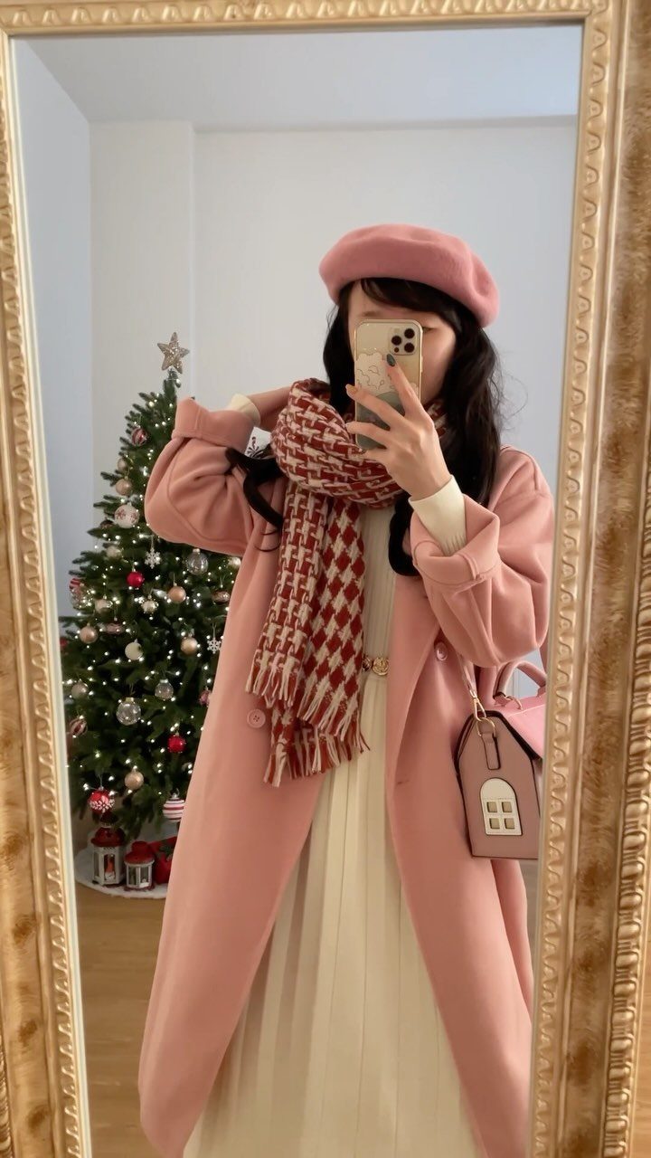 A person wearing a pink coat, patterned scarf, and cream pleated midi dress standing near a Christmas tree.