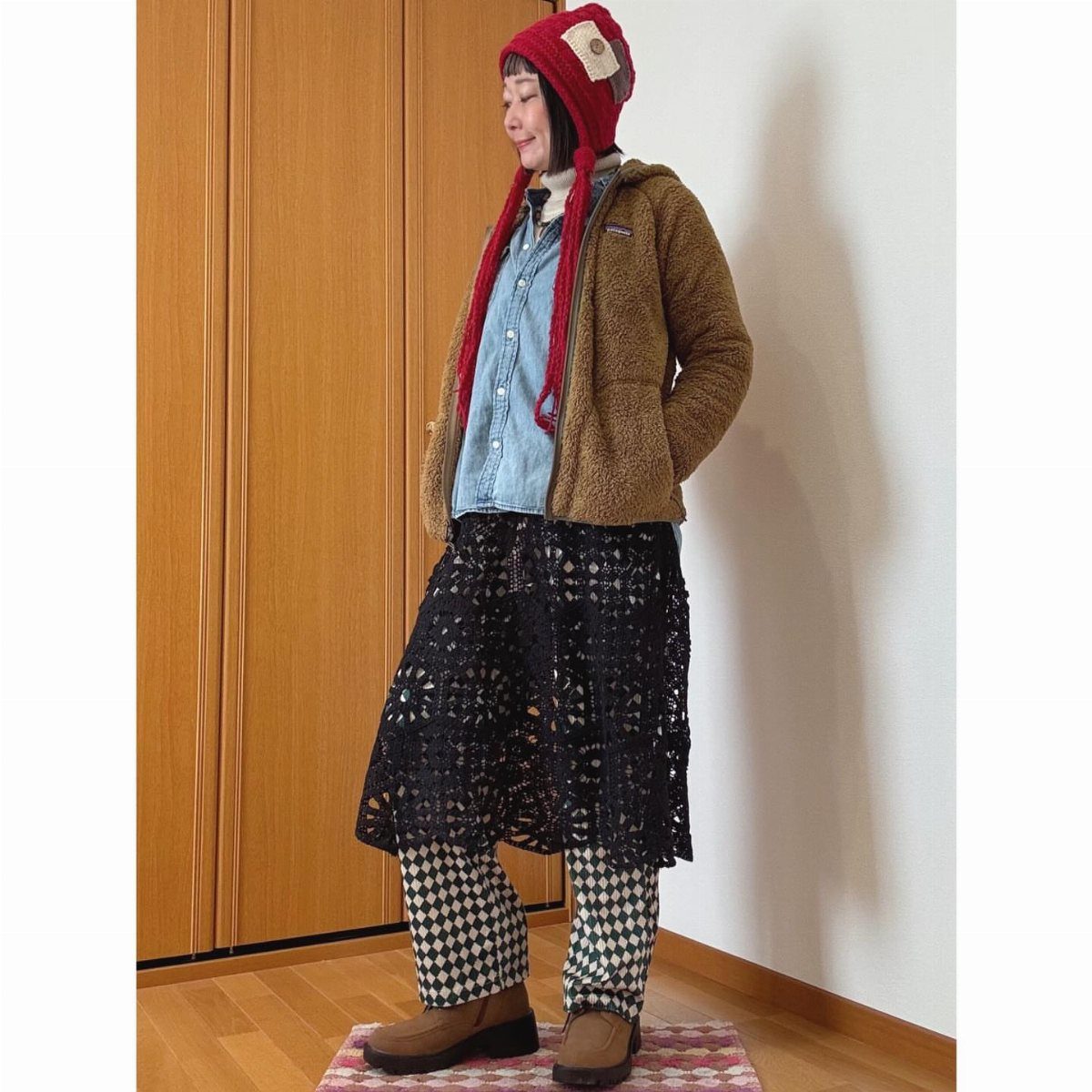 A person wearing a red knit beanie, brown fleece jacket, denim shirt, and black lace skirt.