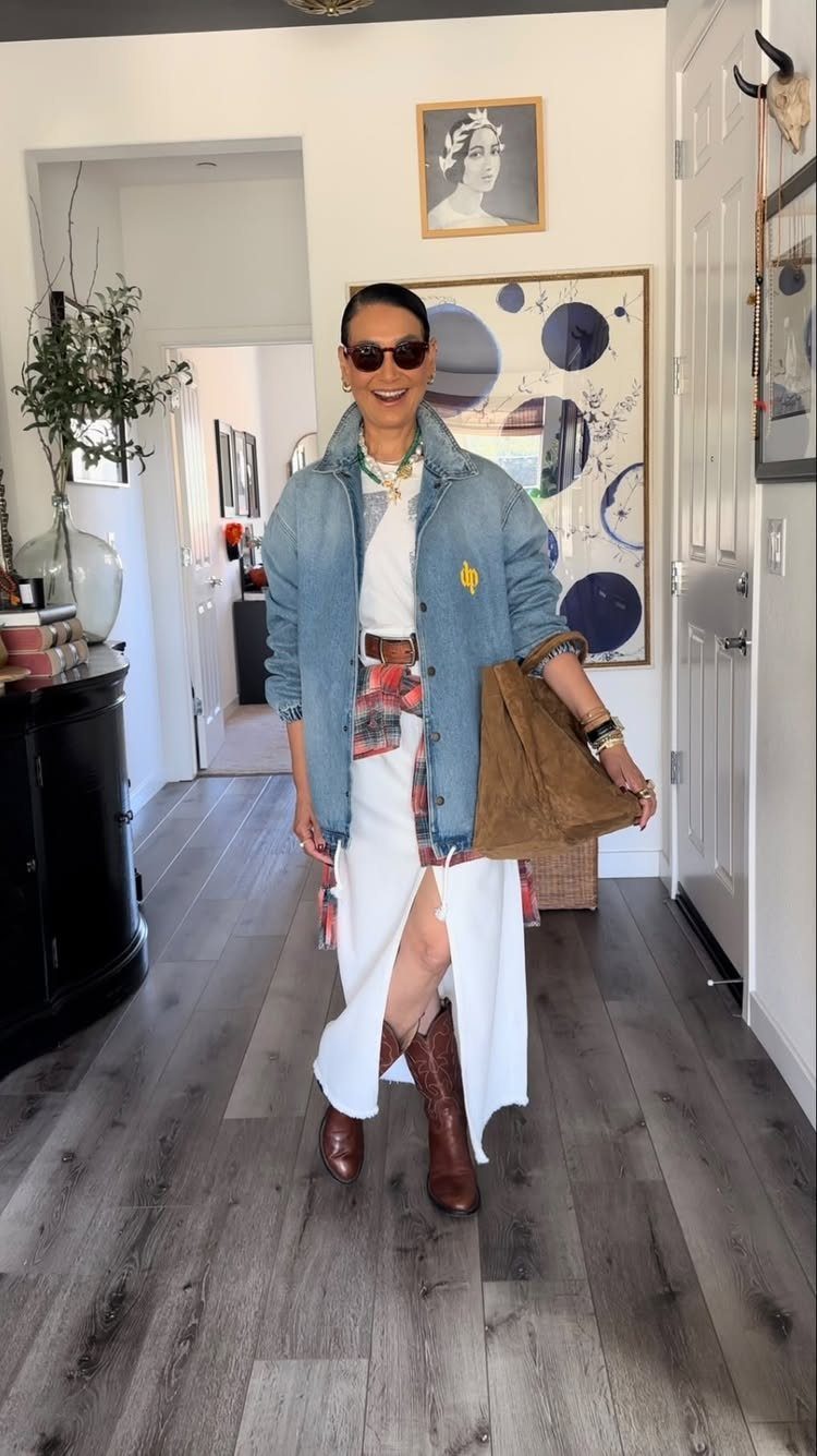 Smiling woman in a denim jacket, white skirt, and brown boots standing in a hallway.