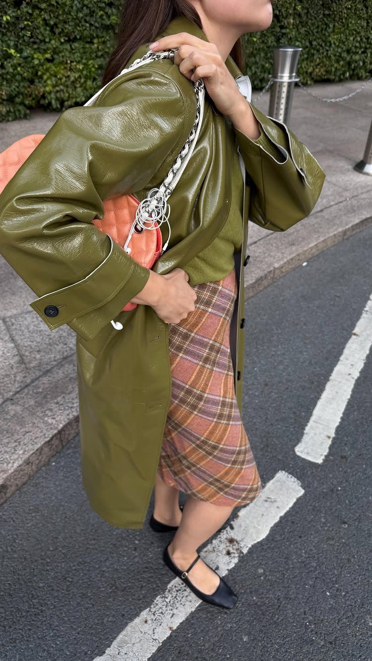 A person wearing an olive green leather coat, plaid skirt, and black ballet flats walking outdoors.