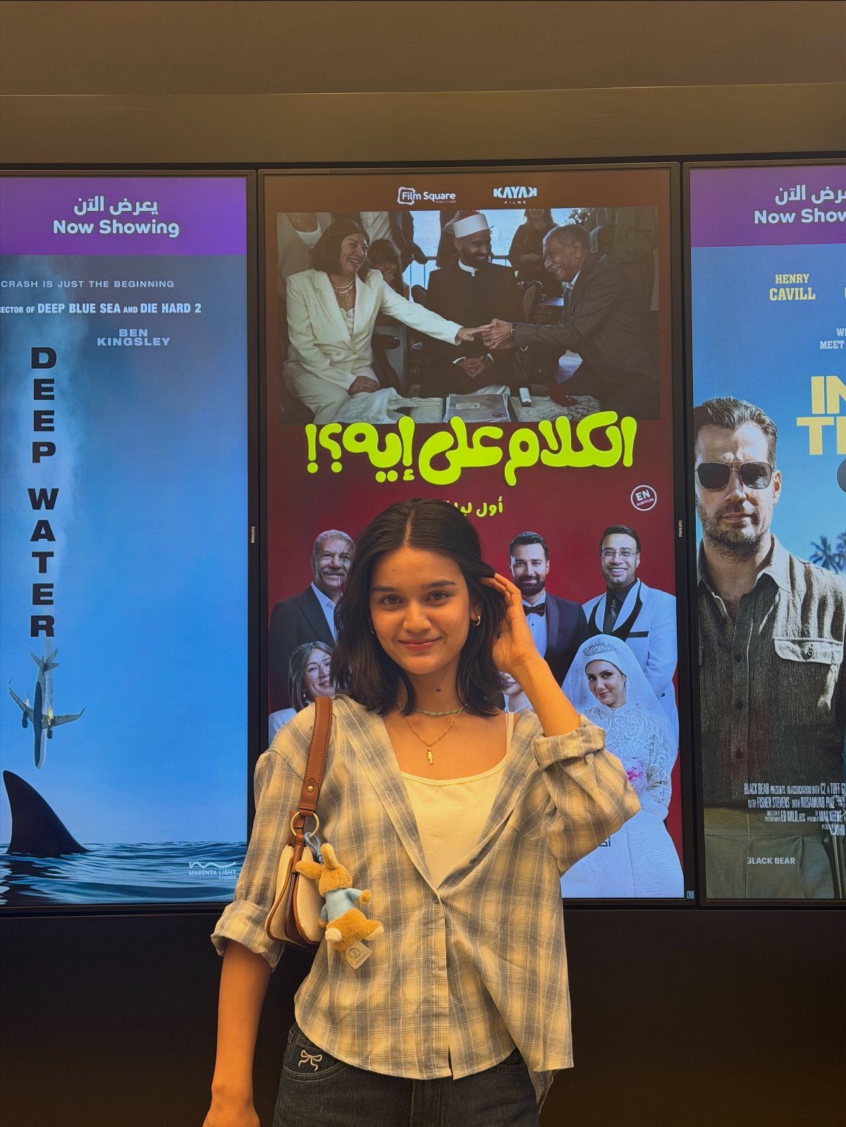 A young woman wearing a plaid flannel shirt and jeans posing in front of movie posters.