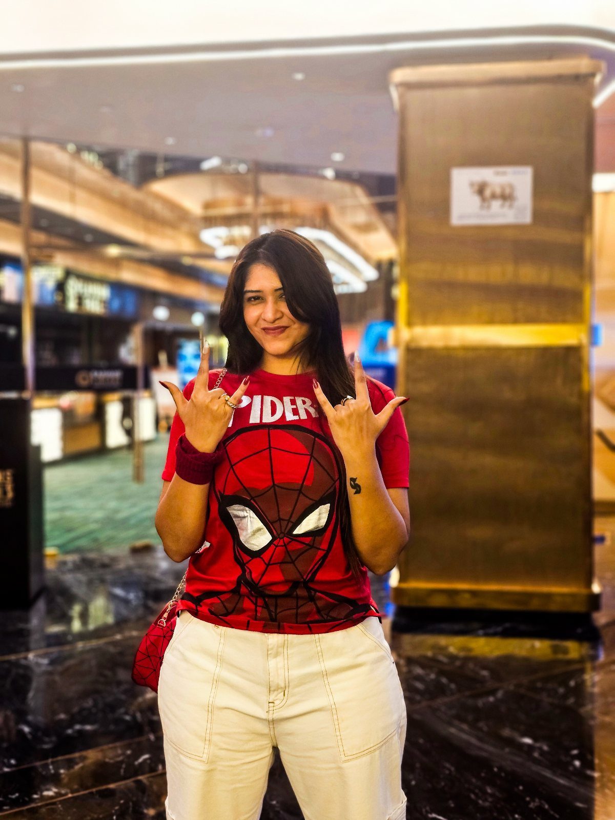 Woman wearing a red Spider-Man graphic t-shirt and white wide-leg pants posing indoors.
