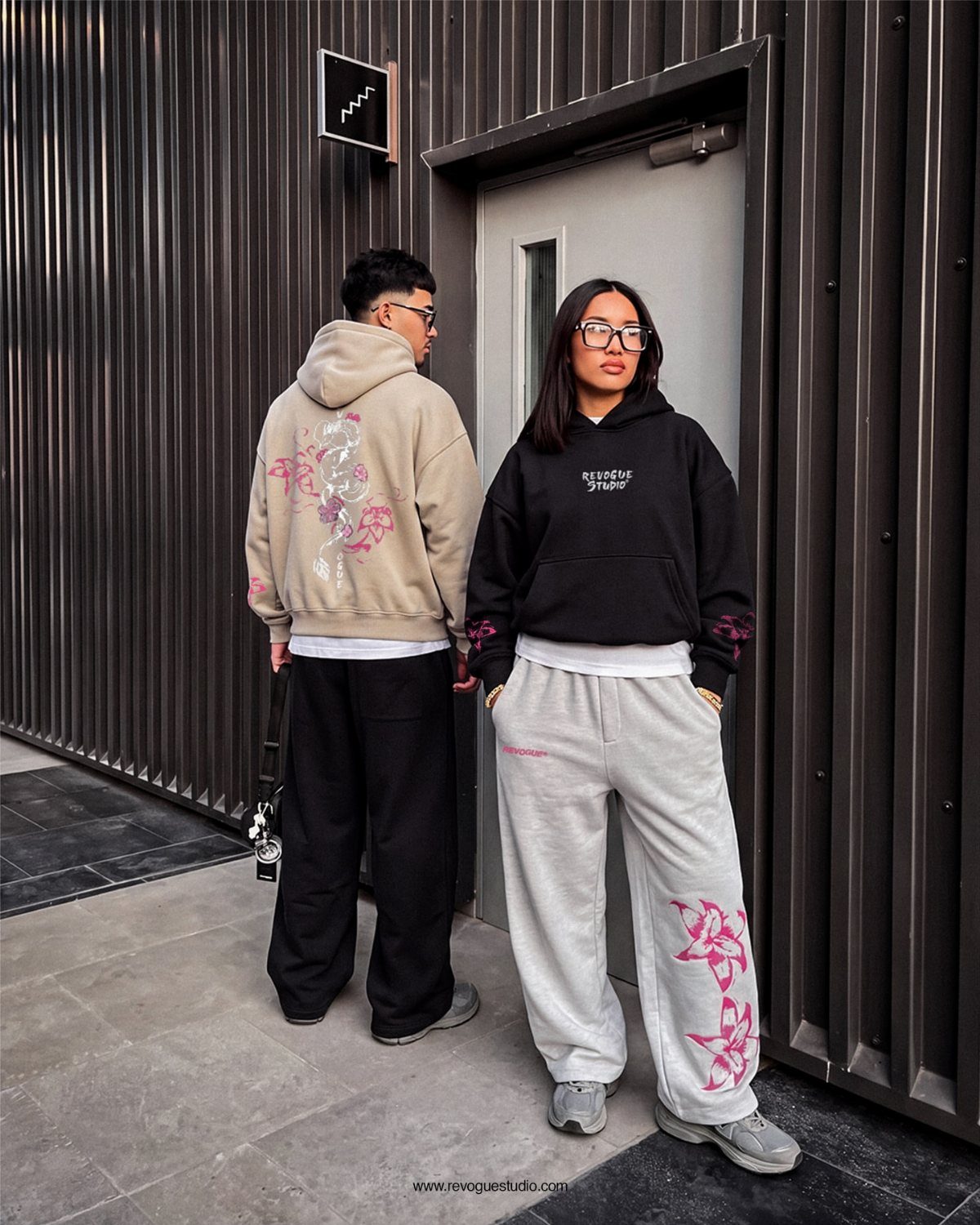 Two people wearing hoodies and sweatpants standing in front of a dark corrugated wall.