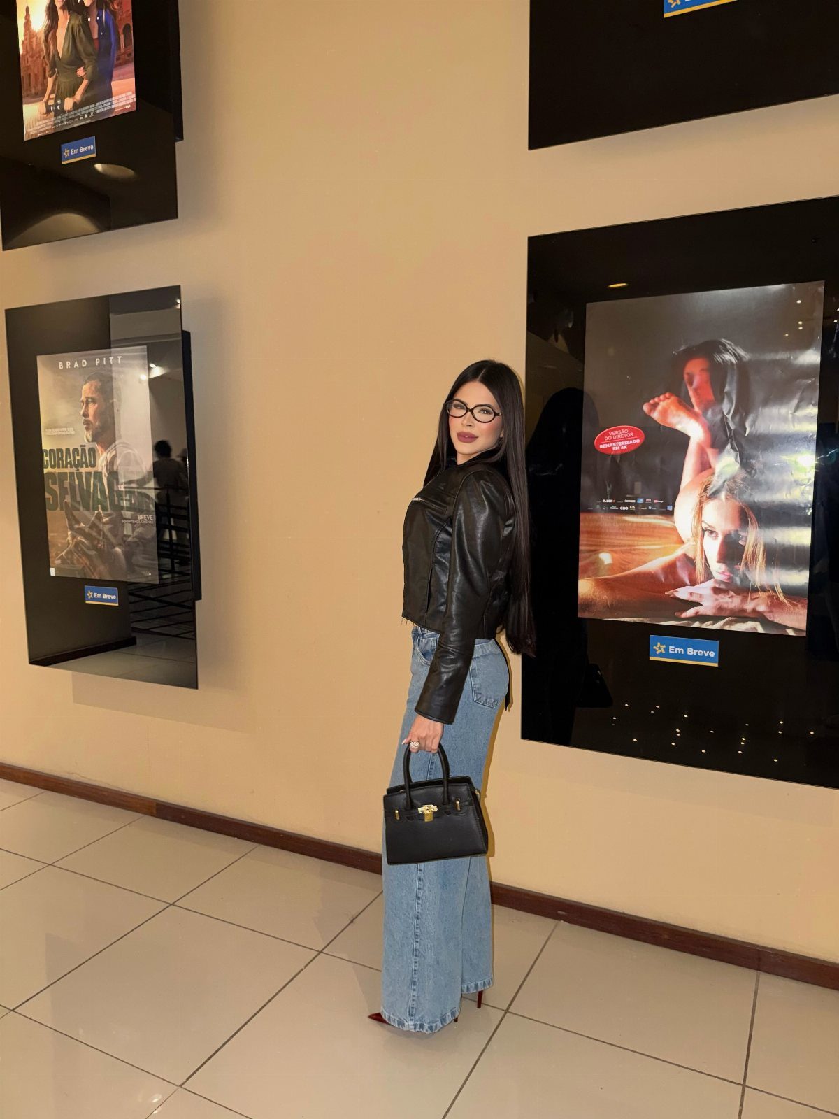 A woman wearing a black leather jacket and wide-leg blue jeans standing near movie posters.