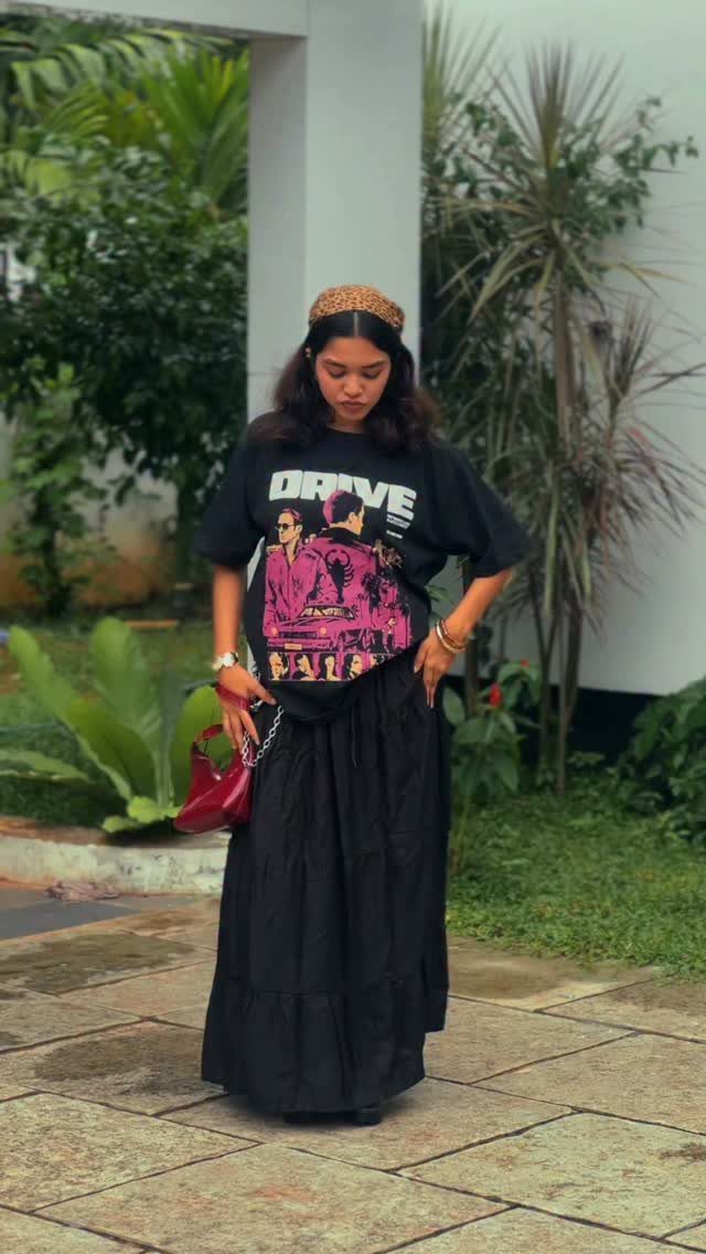 A woman posing in a black graphic t-shirt and a long black maxi skirt outdoors.