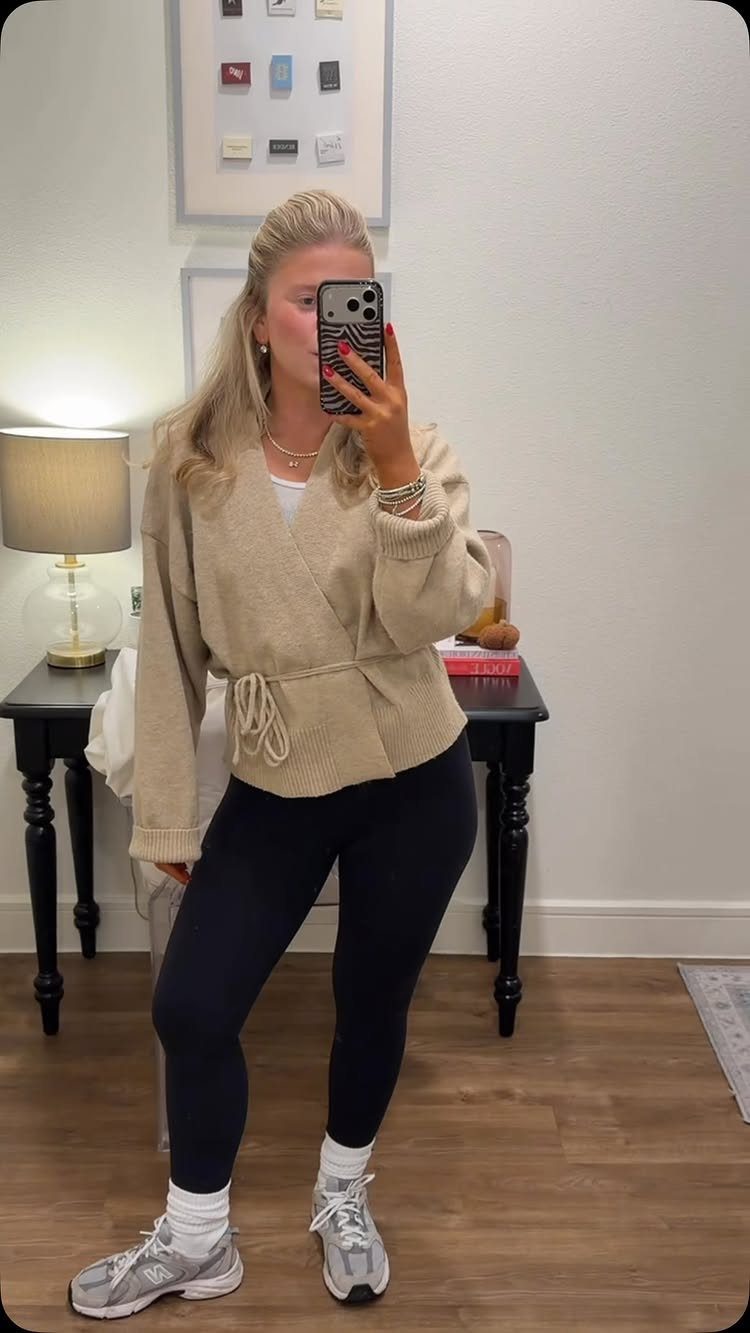 A woman posing in a mirror wearing a beige cardigan, black leggings, and grey sneakers.