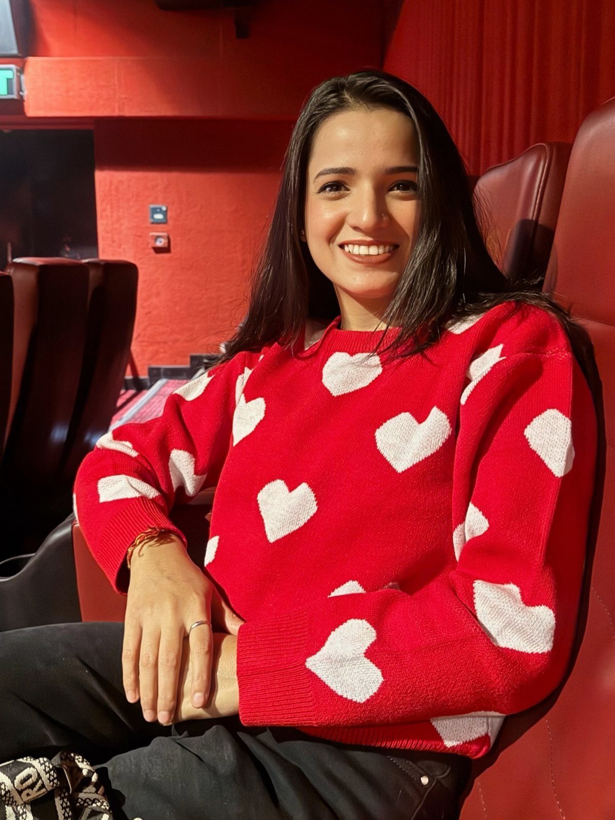 Smiling woman wearing a red sweater with large white hearts while sitting in a theater seat.