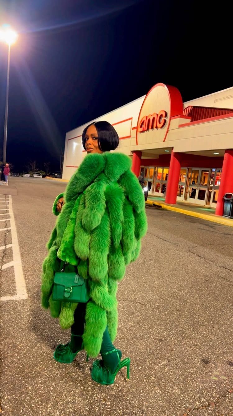 A woman posing in a bright green faux fur coat and matching accessories outside an AMC theater.