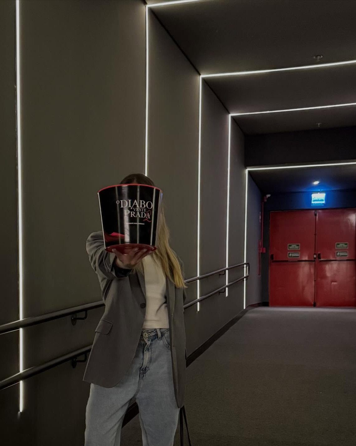 Person in a grey blazer and light blue jeans holding a popcorn bucket in a hallway.