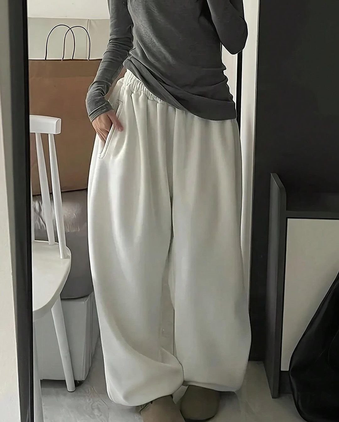 A person wearing a grey long-sleeved shirt and white wide-leg sweatpants standing indoors.