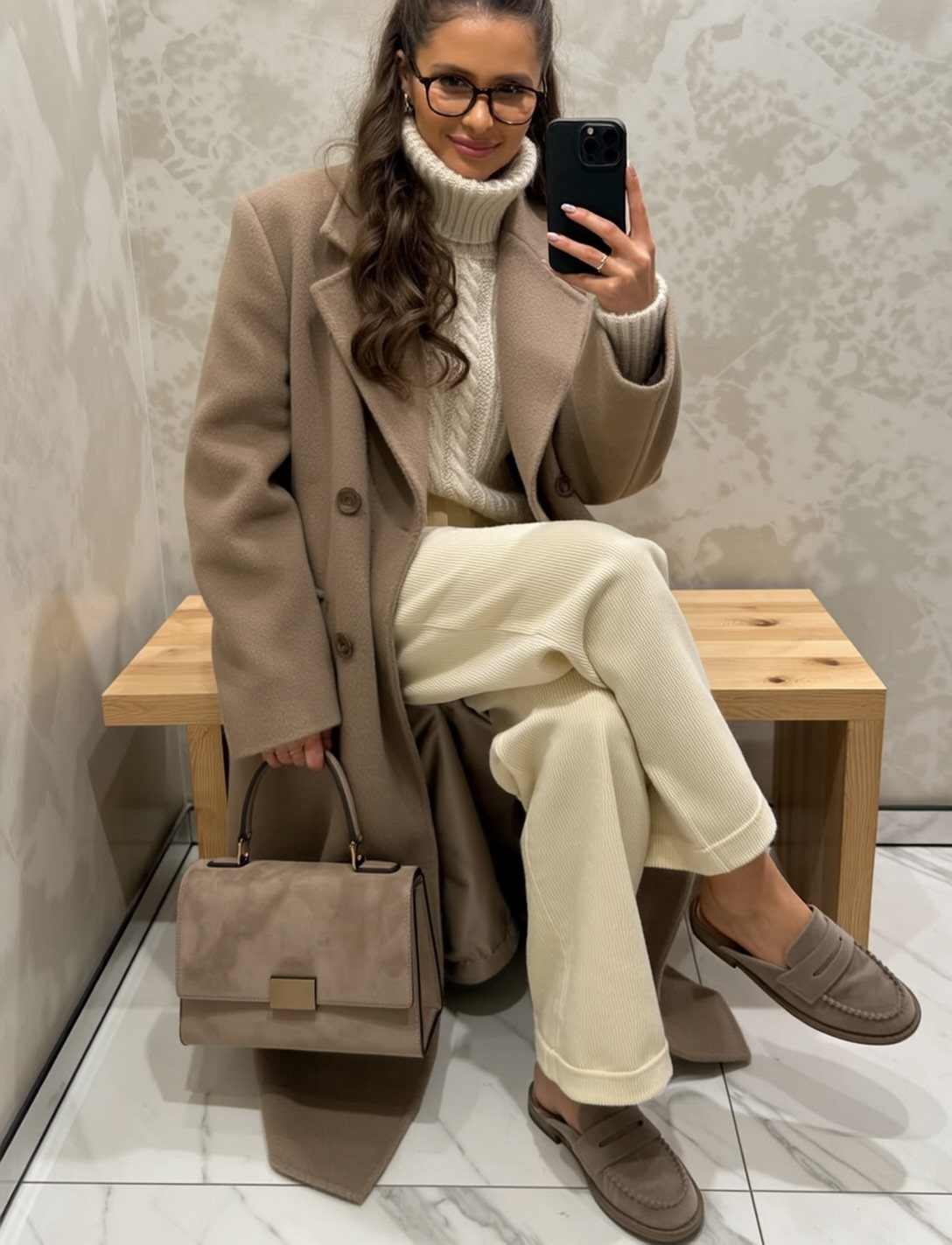 Woman in a tan coat, cream turtleneck, and cream trousers sitting on a wooden bench.