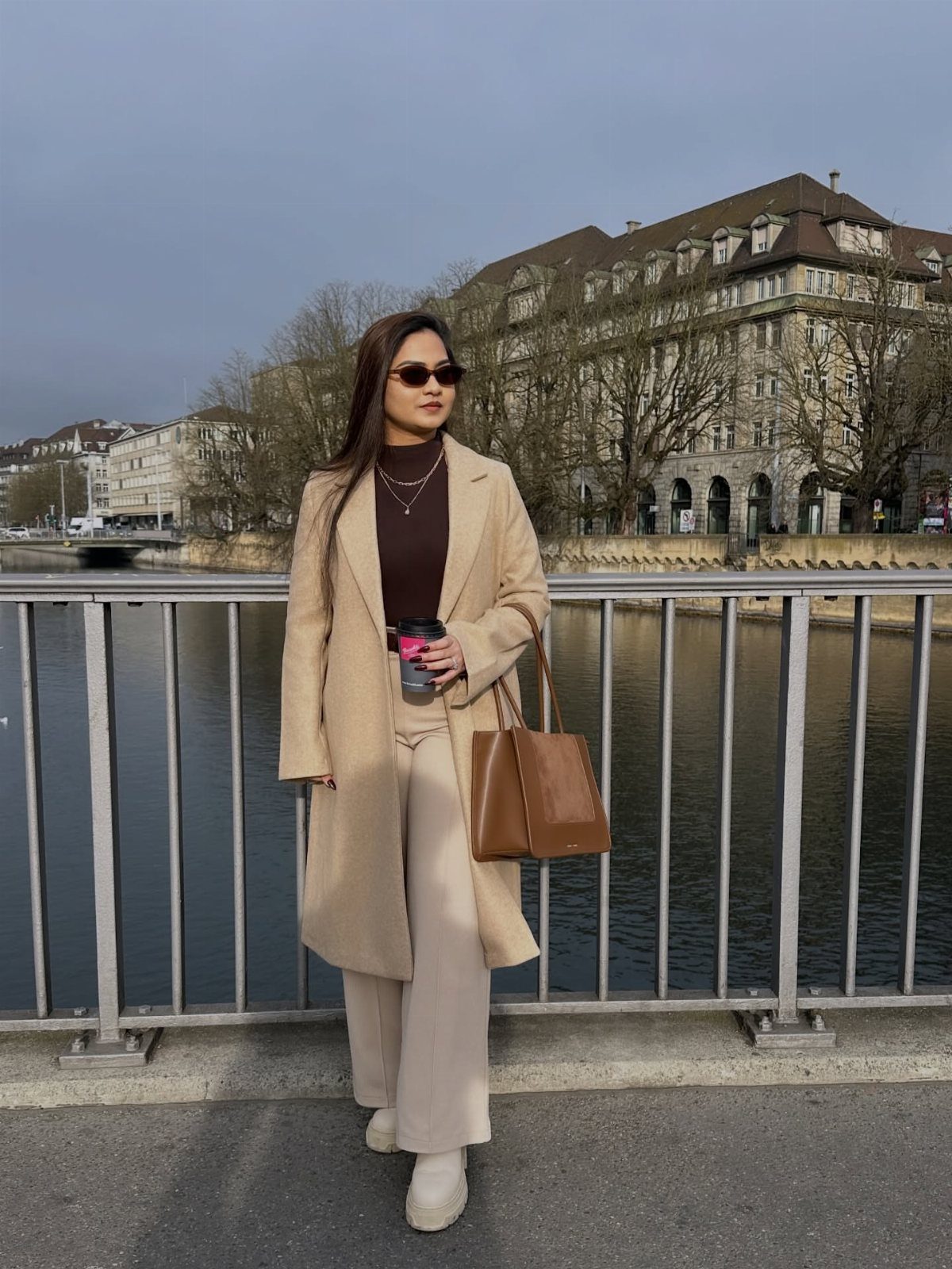 Woman in a tonal neutral winter outfit standing on a bridge by a river.