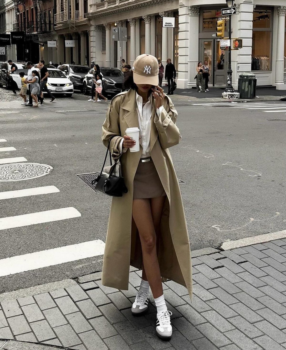 A woman in a tan trench coat, mini skirt, and sneakers standing on a city street.