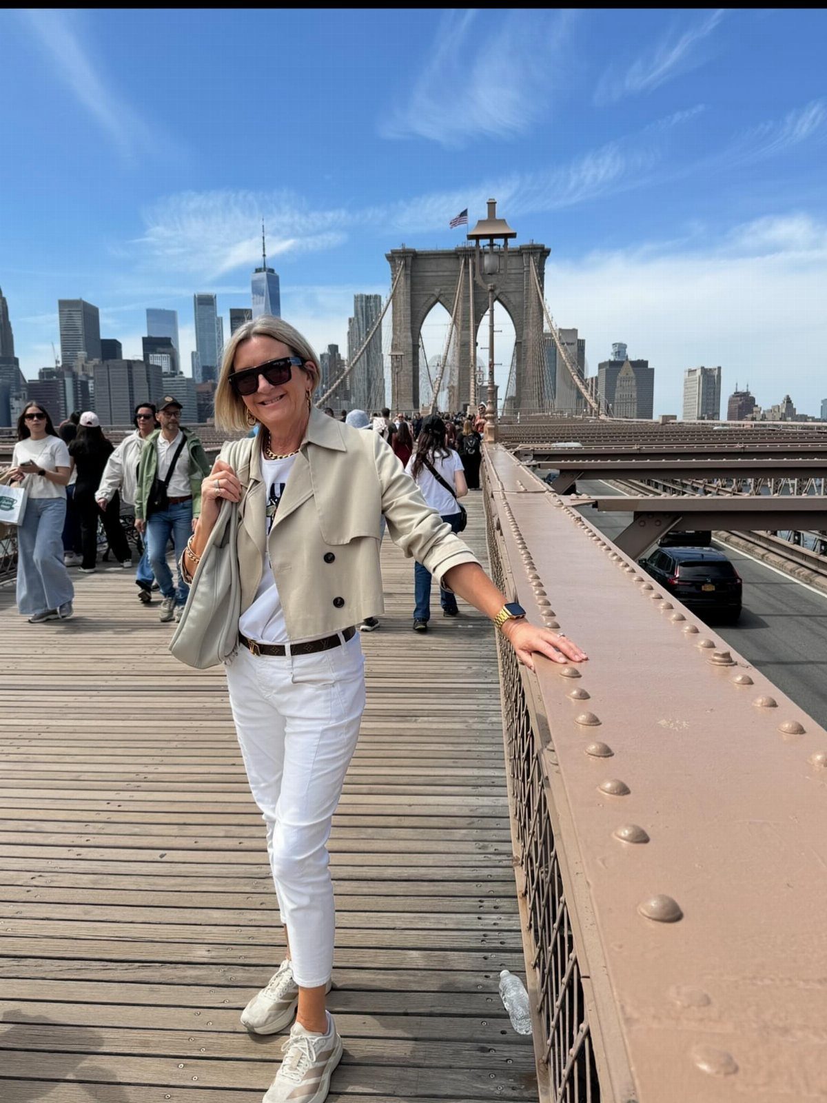 A woman in a beige jacket and white trousers posing on the Brooklyn Bridge.