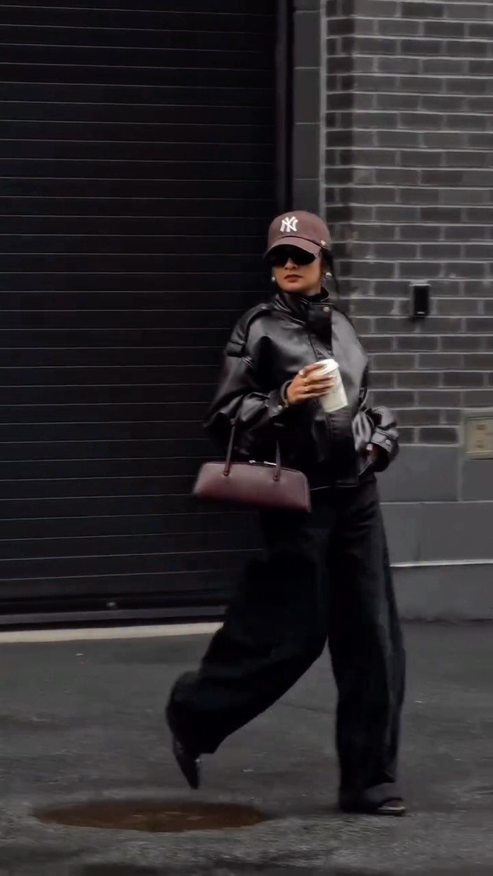 A person in a black leather jacket and wide-leg trousers walking through a city street.