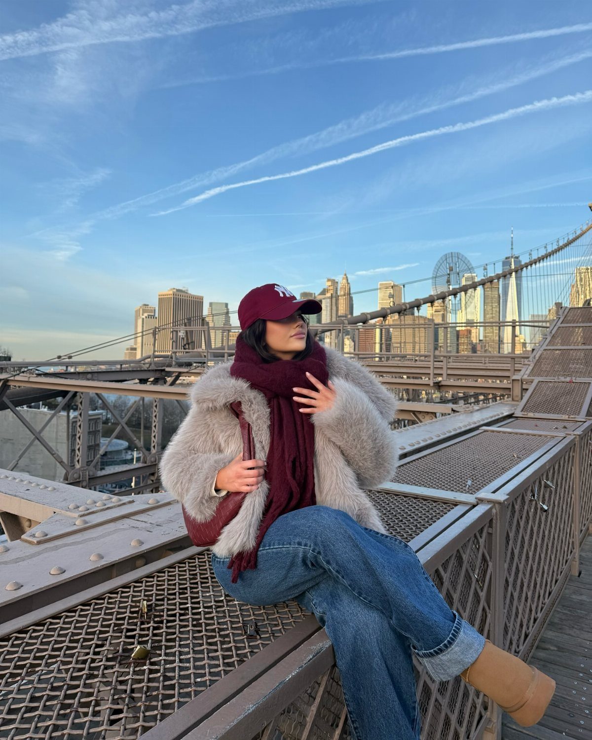 A woman in a faux fur coat and maroon scarf sitting on a bridge overlooking a city skyline.