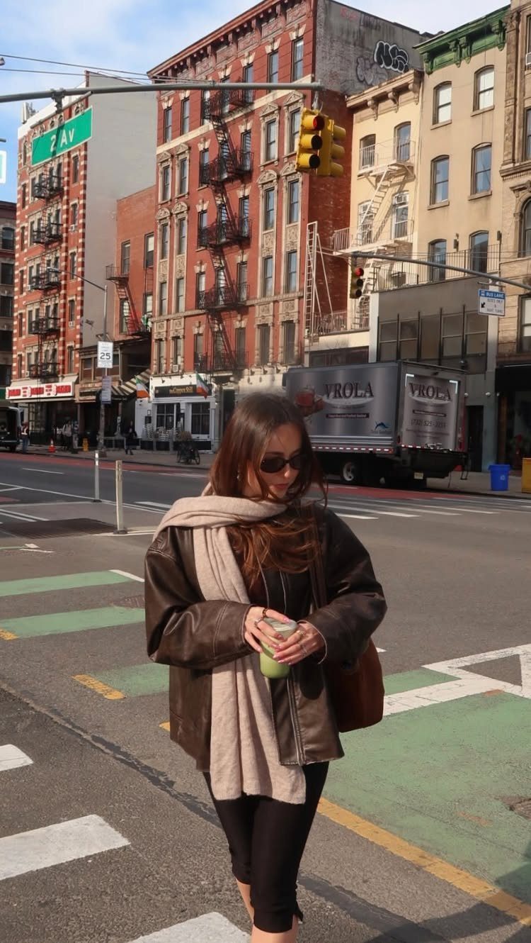 A woman in a leather jacket and large scarf walking across a city street.