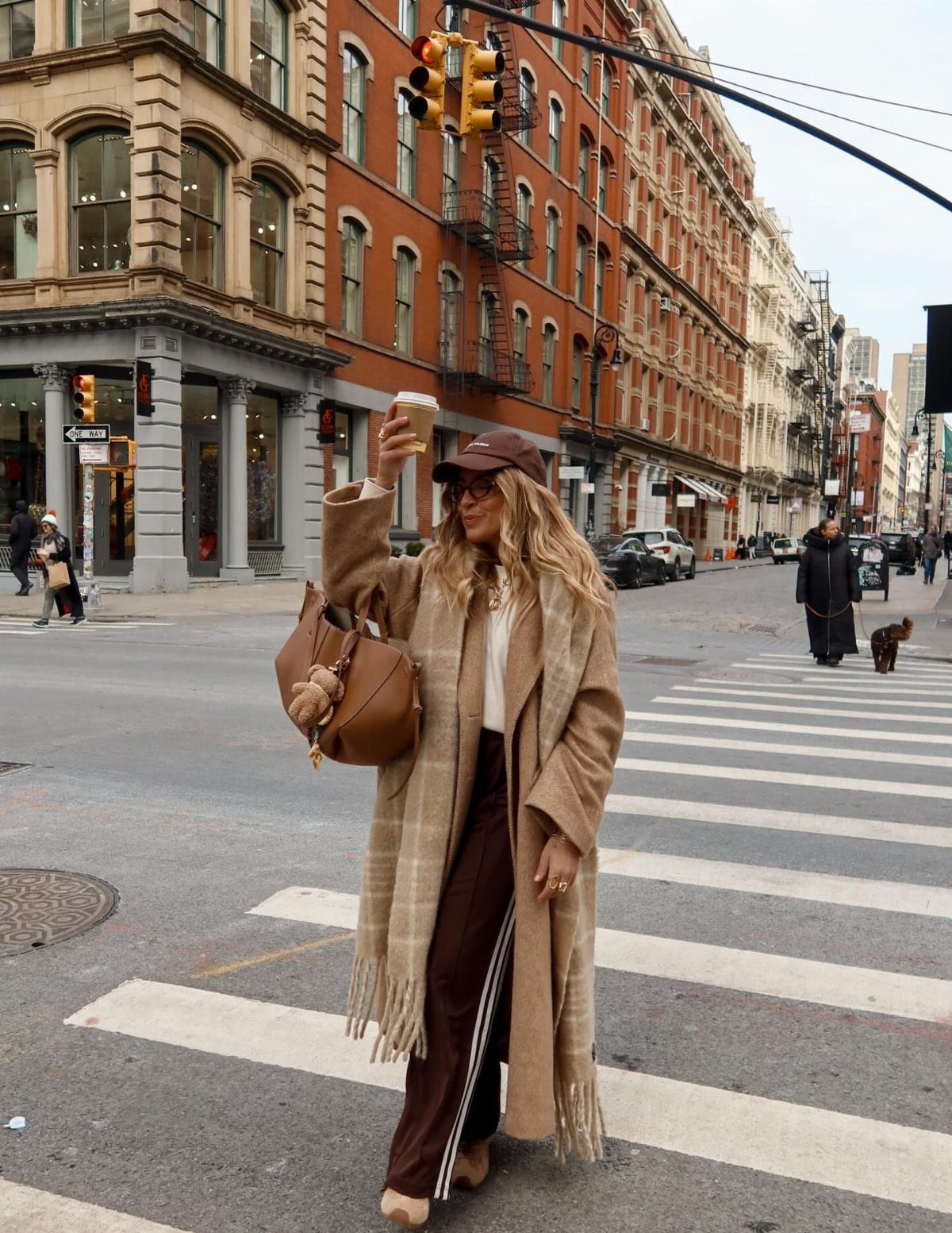A woman in a long tan plaid coat and brown trousers walking across a city street.