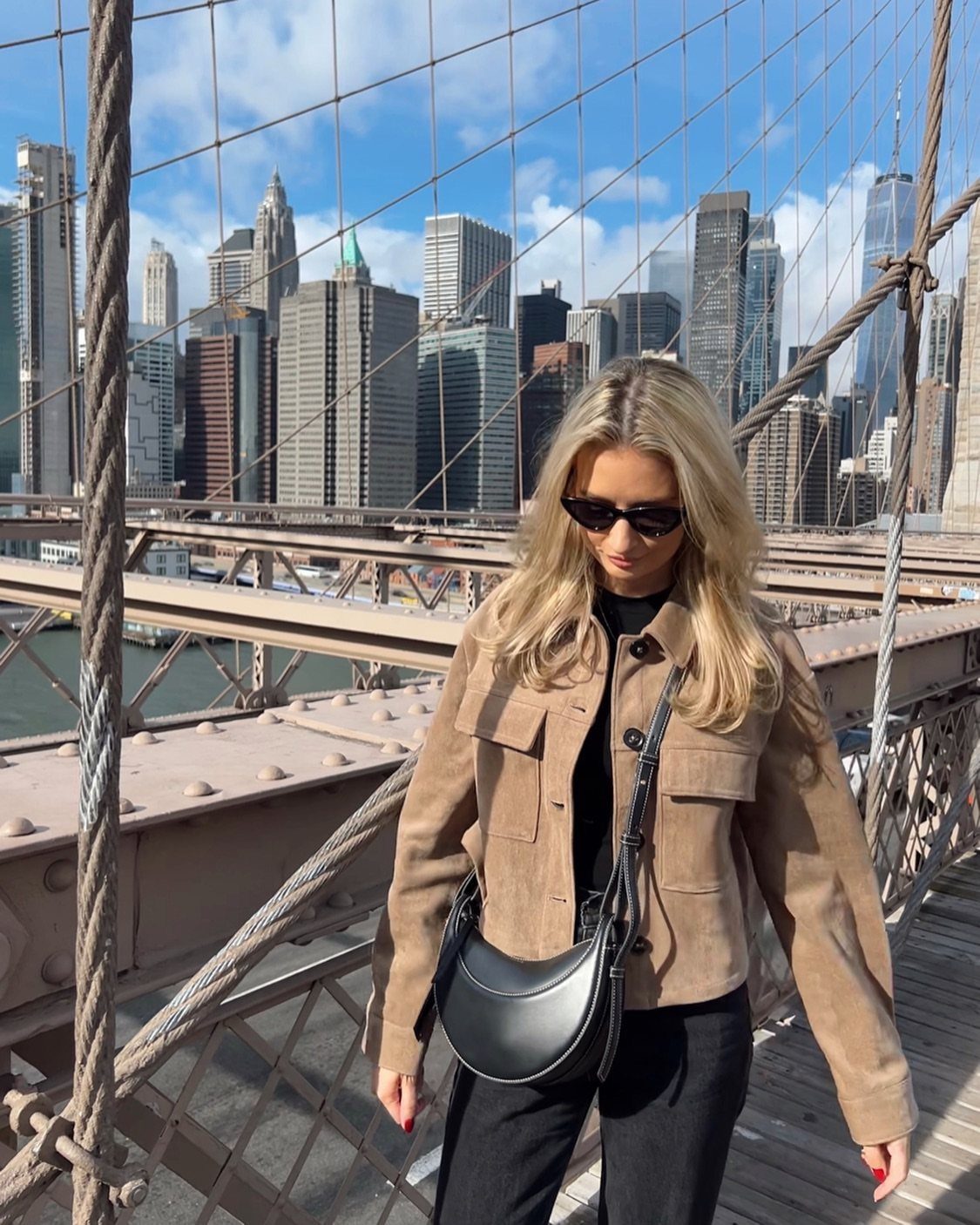 Blonde woman in a tan suede jacket and sunglasses standing on a bridge with city skyscrapers behind her.