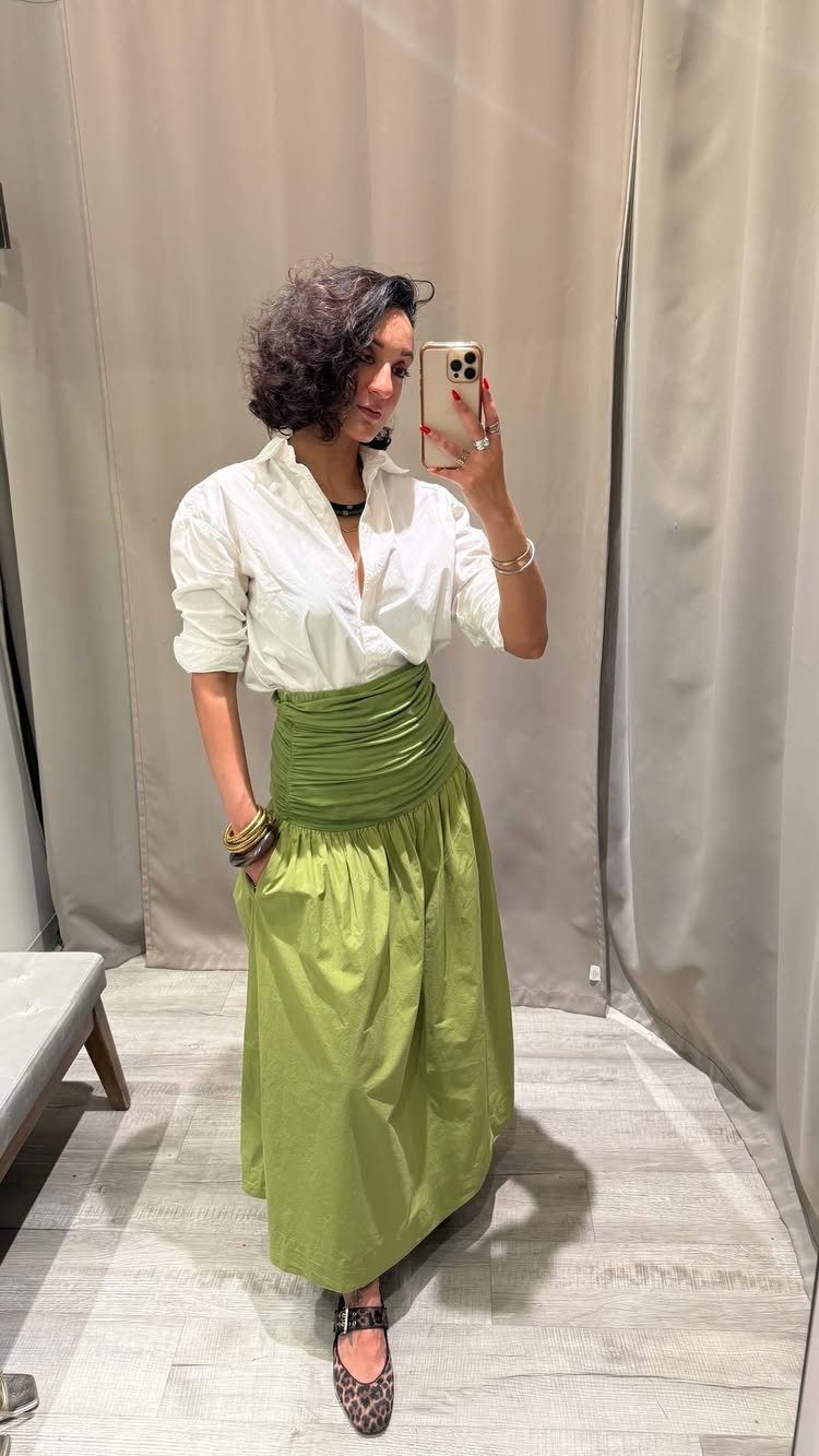 A woman in a white shirt and green maxi skirt taking a mirror selfie.