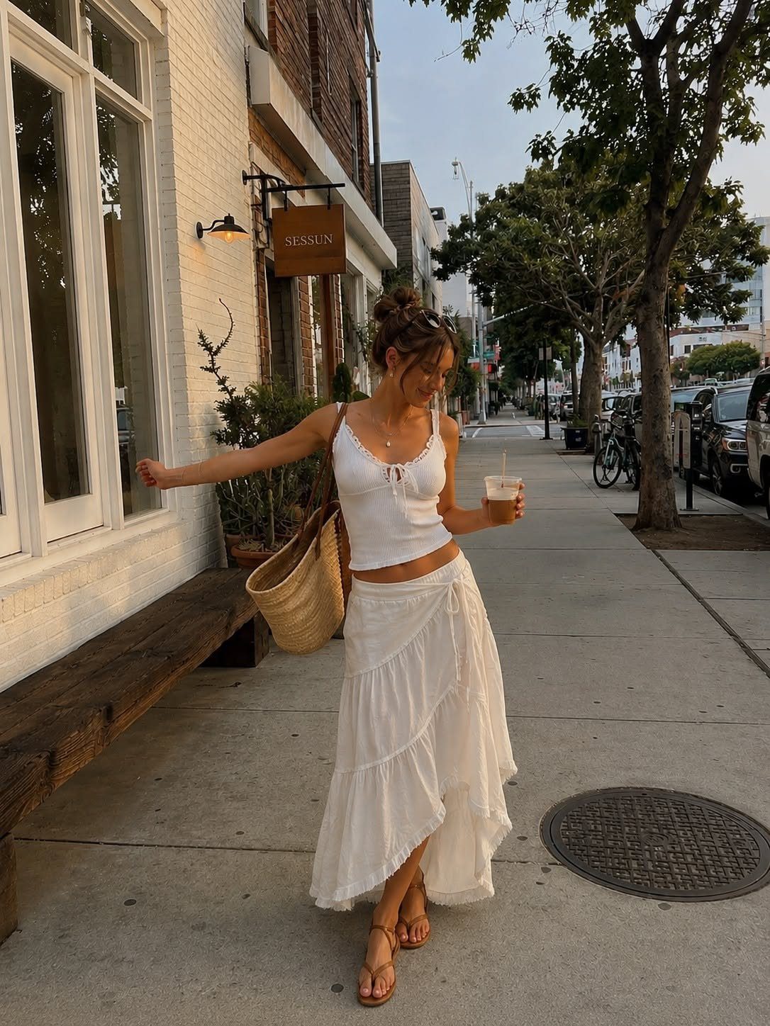 Woman in white summer outfit walking on a city sidewalk carrying a straw bag.